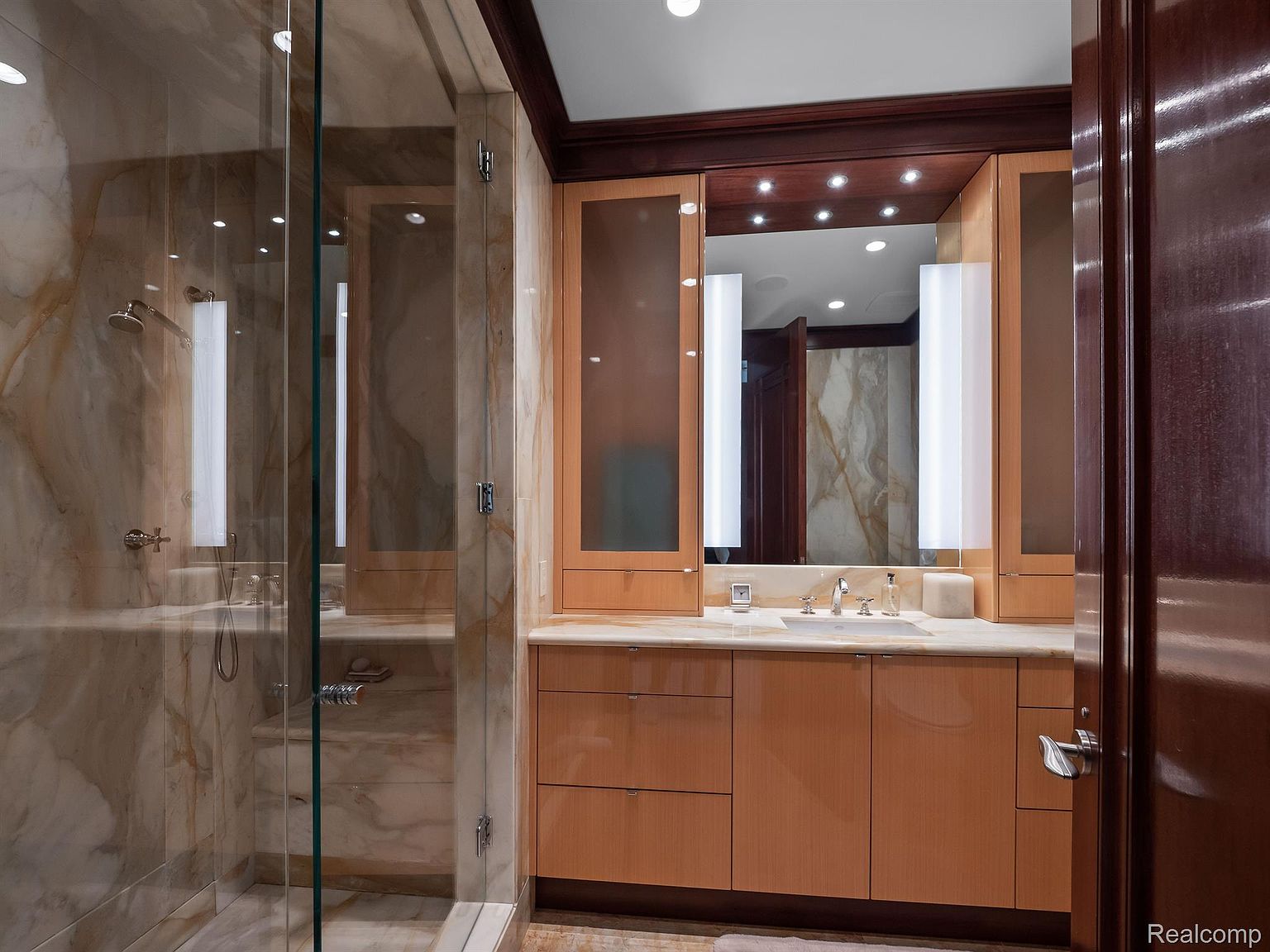 This is a primary bathroom featuring a glass-enclosed shower with marble walls and a vanity with wooden cabinets and a marble countertop. The vanity has a large mirror with integrated lighting, and the overall style is luxurious and modern. A dark wood trim adds a touch of elegance.