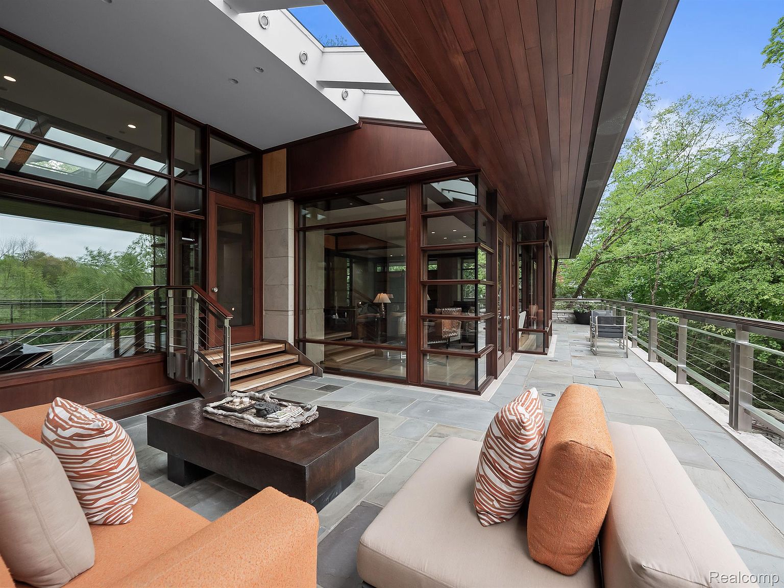 This image showcases a luxurious outdoor patio or deck area. The space features modern architectural elements, including large windows and a wooden overhang. Comfortable outdoor seating with decorative pillows is arranged around a coffee table, creating an inviting atmosphere for relaxation and entertainment.