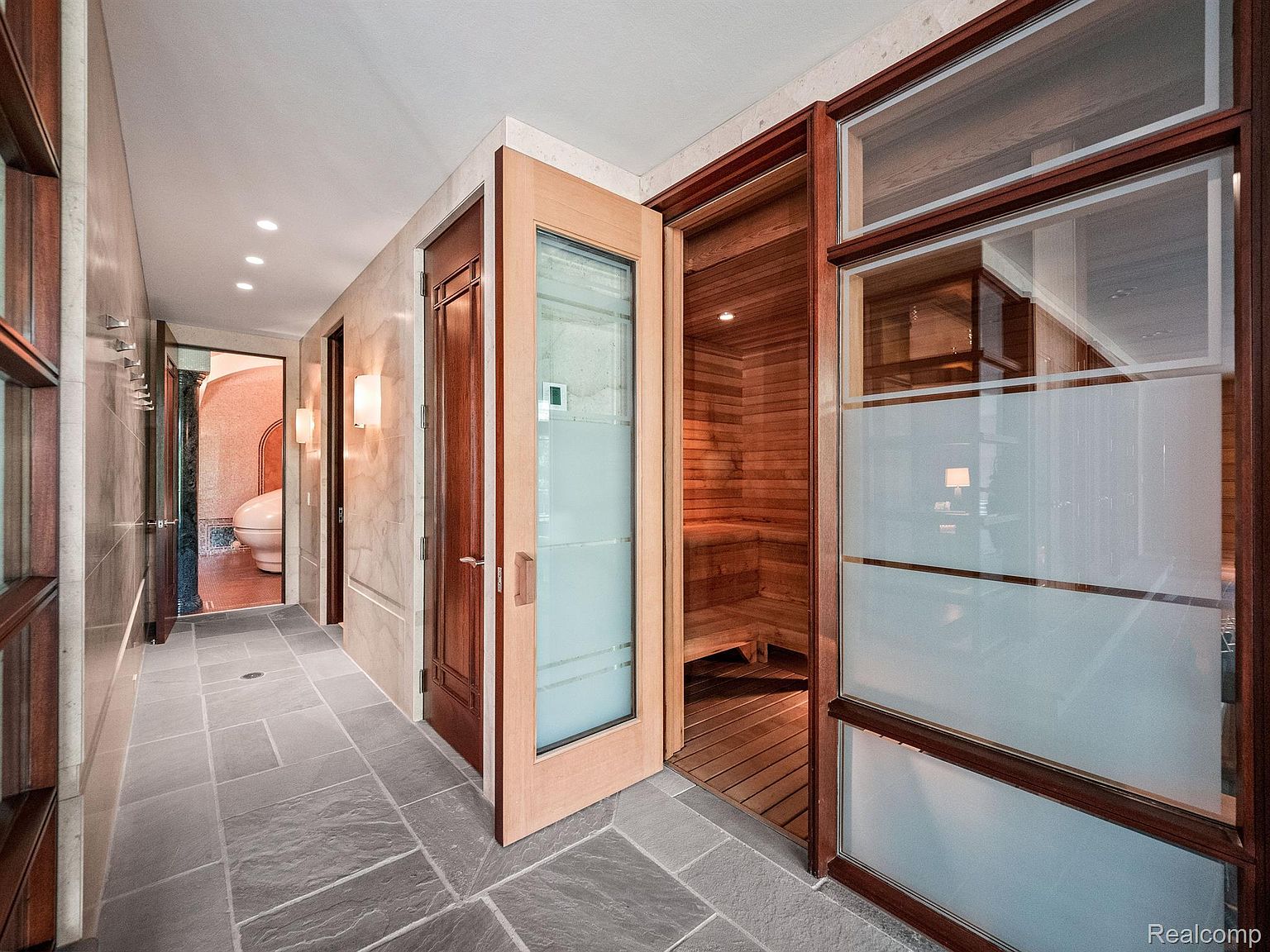 This interior shot showcases a hallway with stone tile flooring, leading to a bathroom and a sauna. The hallway features recessed lighting and wall sconces, creating a warm ambiance. The sauna is visible through a glass door, highlighting its wood paneling and bench seating.