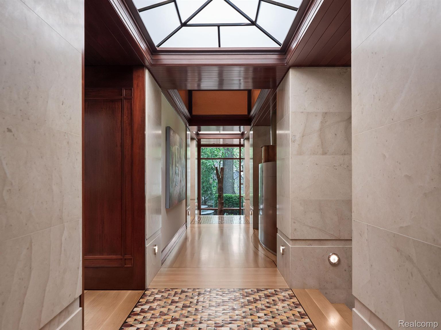 This interior shot showcases a luxurious hallway with a unique geometric tile accent at the entrance. The hallway features light-colored stone walls, rich wood trim, and a striking skylight that floods the space with natural light. The design emphasizes clean lines and high-end materials, creating an elegant and inviting atmosphere.