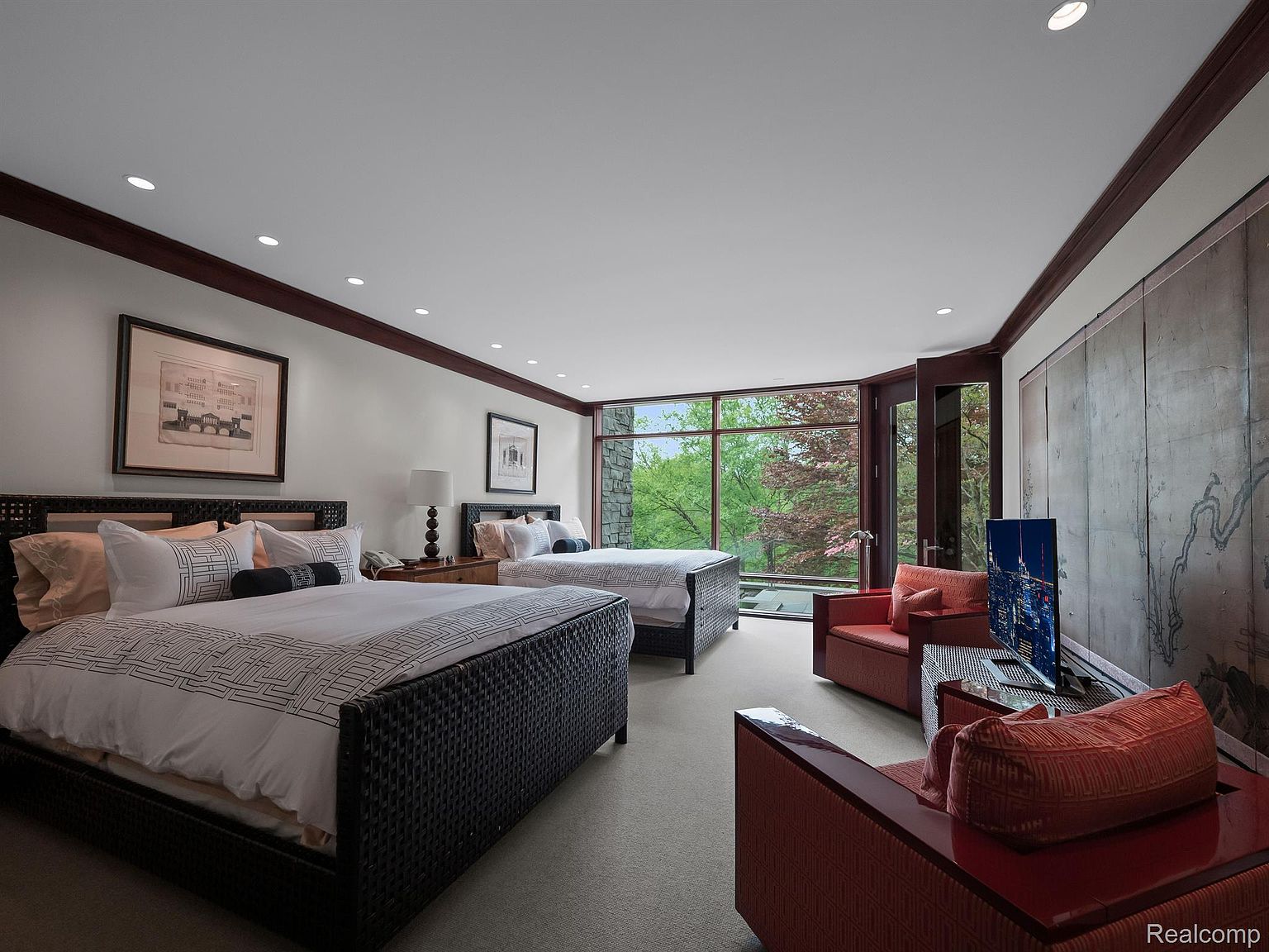 This primary bedroom features two beds with dark wood frames and white linens, complemented by matching nightstands and lamps. A large window offers a view of the outdoors, while a seating area with red armchairs and a television provides a comfortable space for relaxation. The room is spacious and well-lit, creating a luxurious and inviting atmosphere.