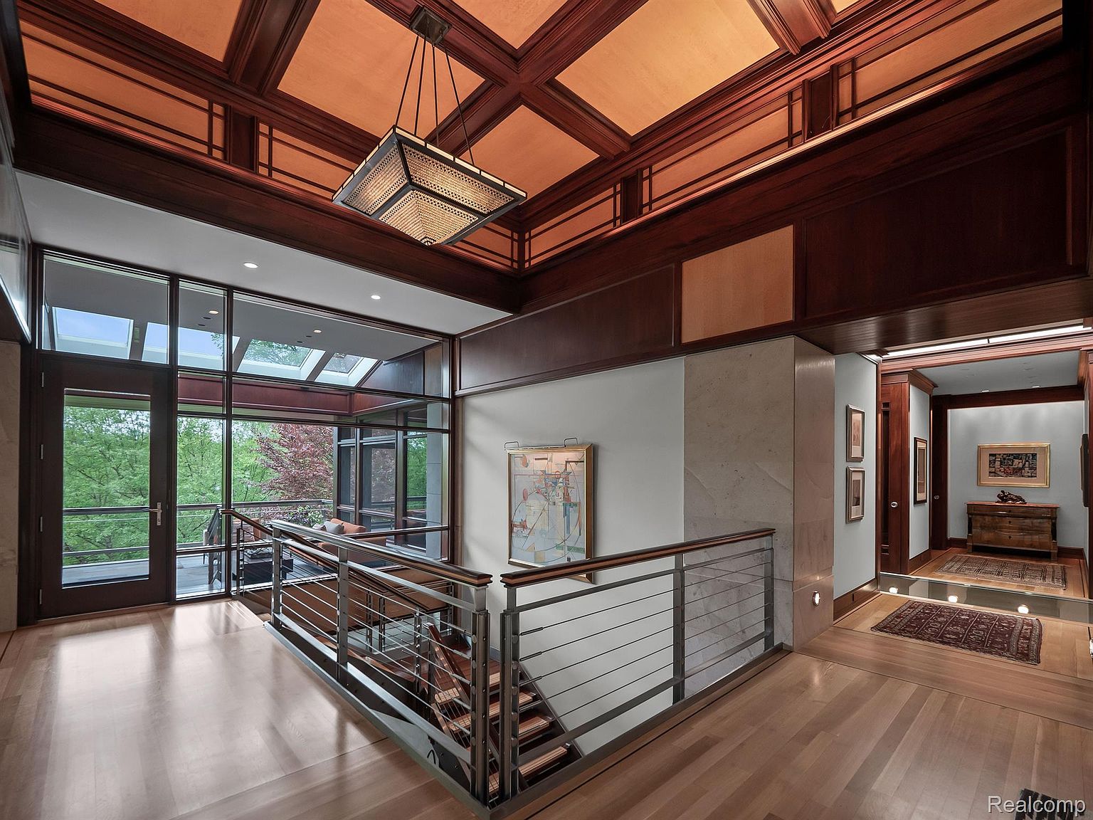 This interior shot showcases a luxurious hallway and staircase. The space features rich wood paneling on the ceiling, a modern chandelier, and a large window offering natural light and views of the outdoors. A stainless steel railing complements the wooden staircase, leading to a lower level, while a doorway leads to another room with a rug and wooden furniture.