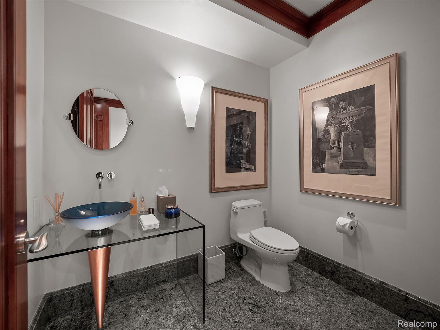 This is a stylish guest bathroom featuring a unique glass countertop with a blue vessel sink and a copper-colored pedestal. The walls are painted a neutral gray, complemented by framed artwork and modern sconce lighting. The flooring is a dark, speckled granite, adding a touch of luxury to the space.