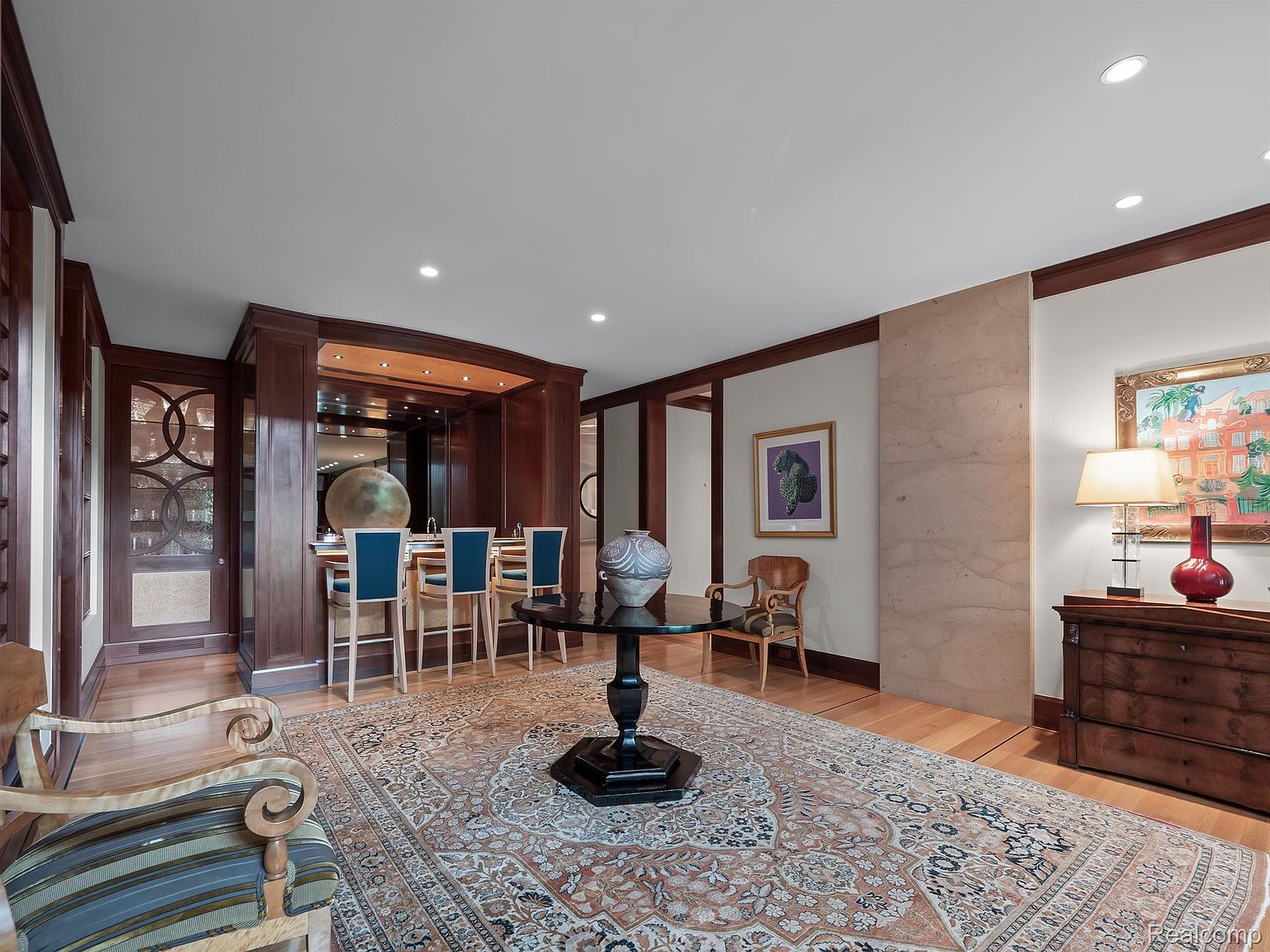 This interior shot showcases a sophisticated living room with rich wood accents and a large, ornate rug. A bar area with seating is visible in the background, along with decorative elements like a globe and a large vase on a center table. The room exudes a sense of classic elegance and comfort, perfect for entertaining or relaxing.