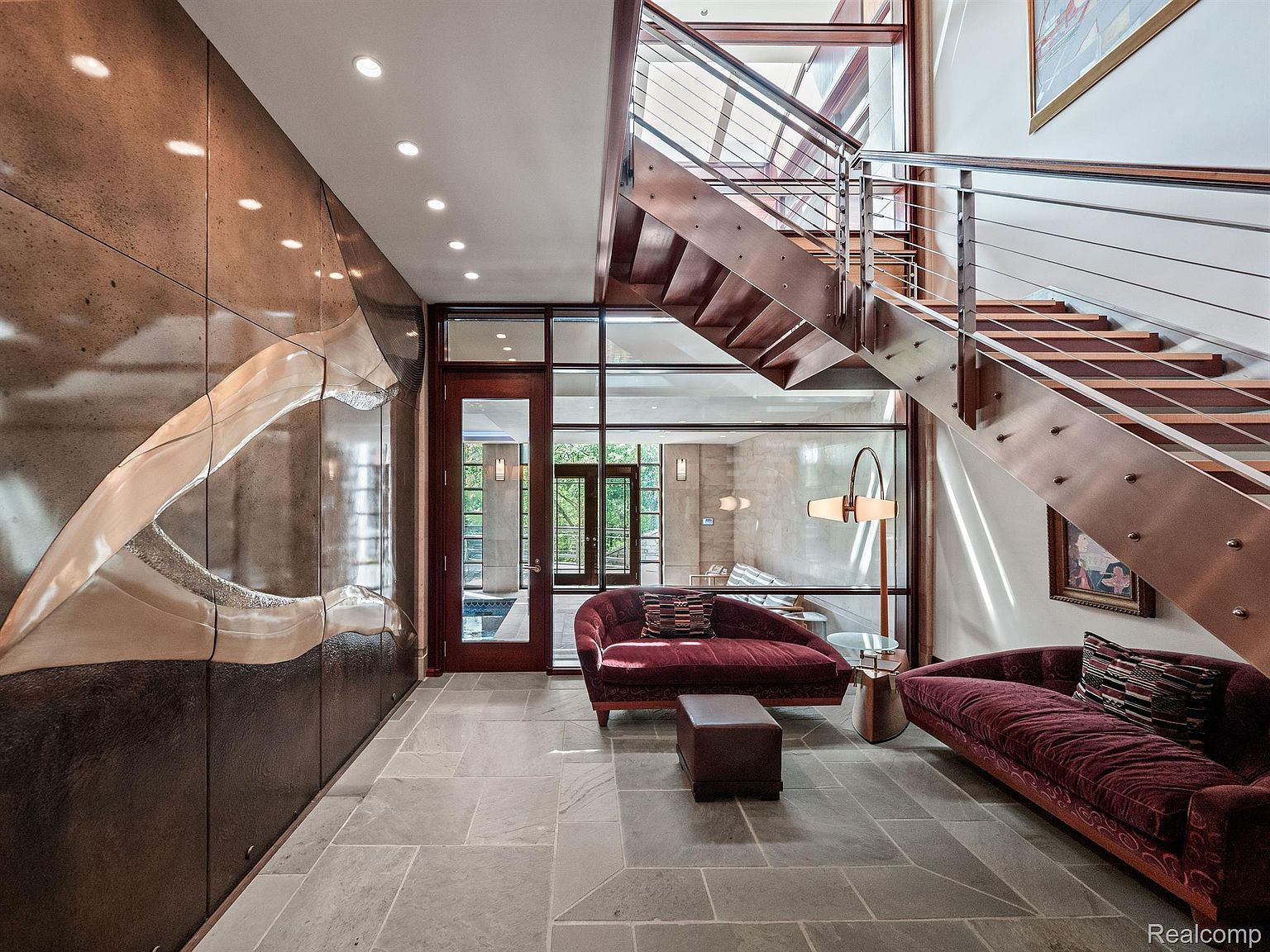This interior shot showcases a modern hallway with a striking architectural wall feature and a floating staircase with stainless steel railings. The space is furnished with stylish maroon sofas and a small ottoman, creating a welcoming and sophisticated atmosphere. Large windows and a glass door provide ample natural light, enhancing the contemporary design.