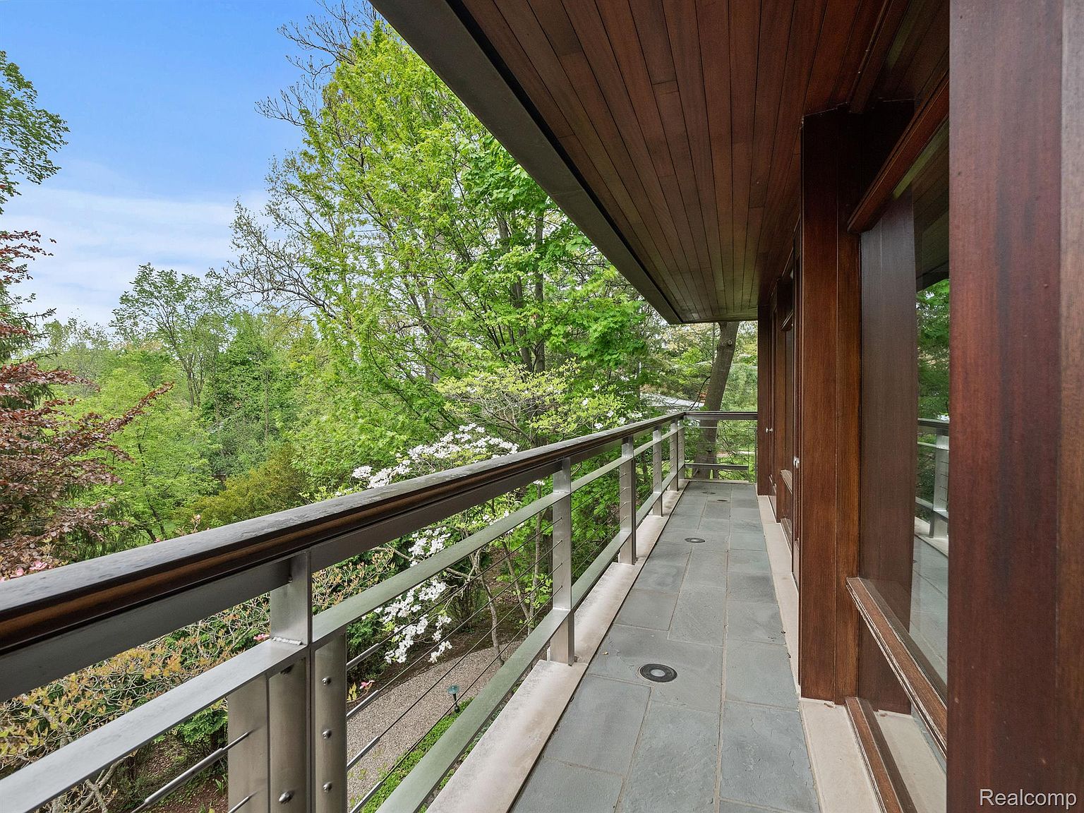 This image showcases a modern balcony with a sleek metal railing and a wooden ceiling. The flooring consists of gray stone tiles, and the balcony offers a view of lush greenery and trees. The overall impression is one of contemporary design and outdoor living space.