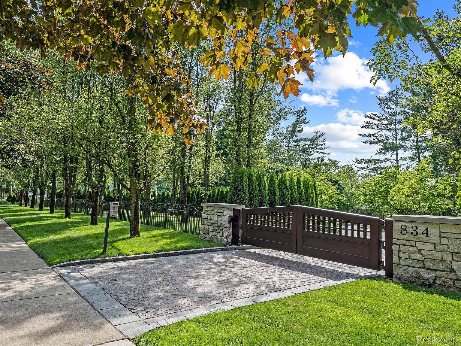 This is a front exterior view of a property featuring a gated entrance with stone pillars and the house number '834' displayed. The driveway is paved with a circular pattern, and a well-maintained lawn extends to the sidewalk. Mature trees line the street, providing shade and privacy, creating an inviting and upscale curb appeal.