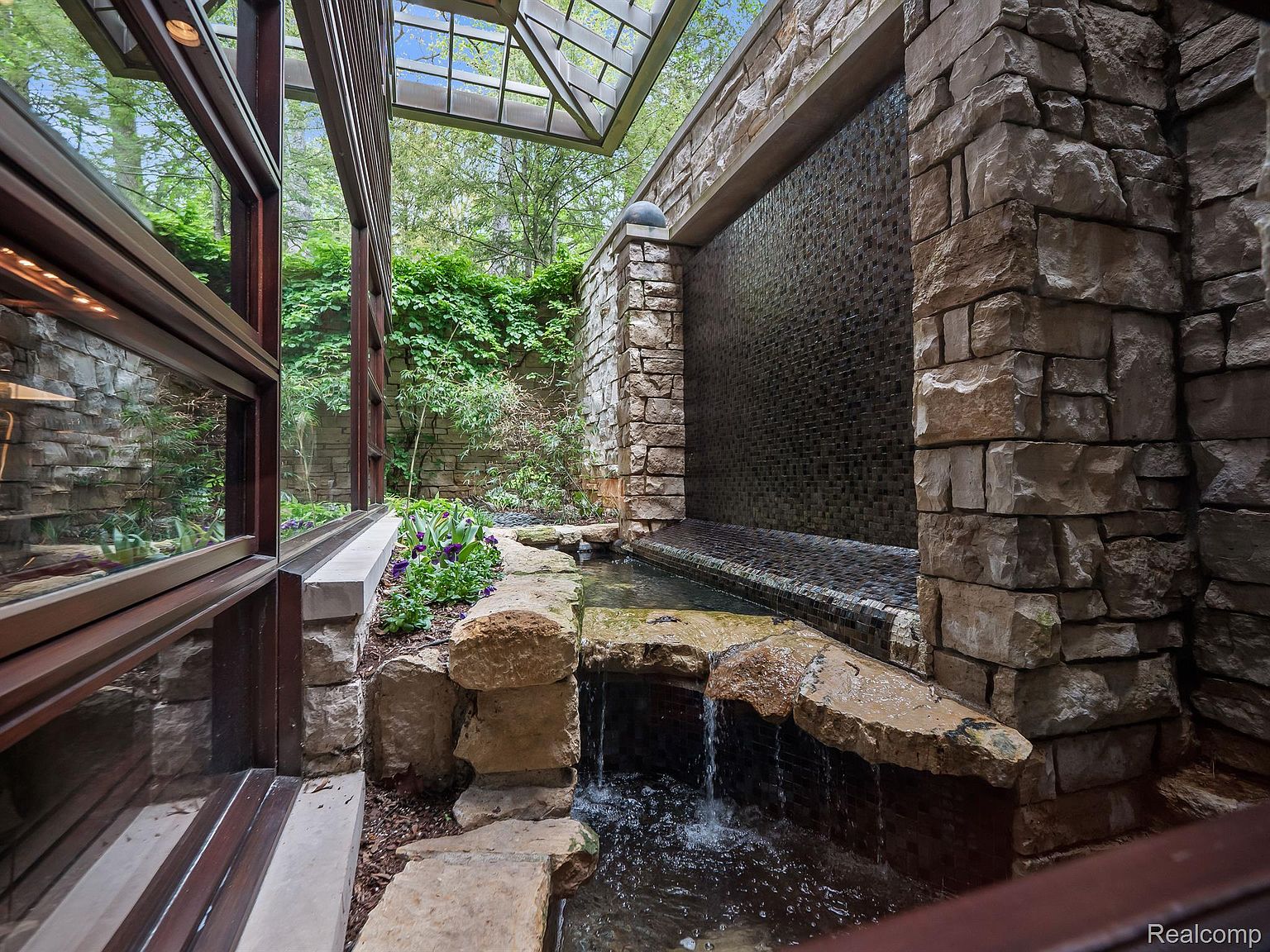 This image showcases a unique architectural feature: an indoor waterfall and pond integrated into the building's design. The waterfall cascades down a textured wall into a stone-lined pond, surrounded by lush greenery and natural stone elements. A glass ceiling allows natural light to filter in, creating a serene and luxurious ambiance.