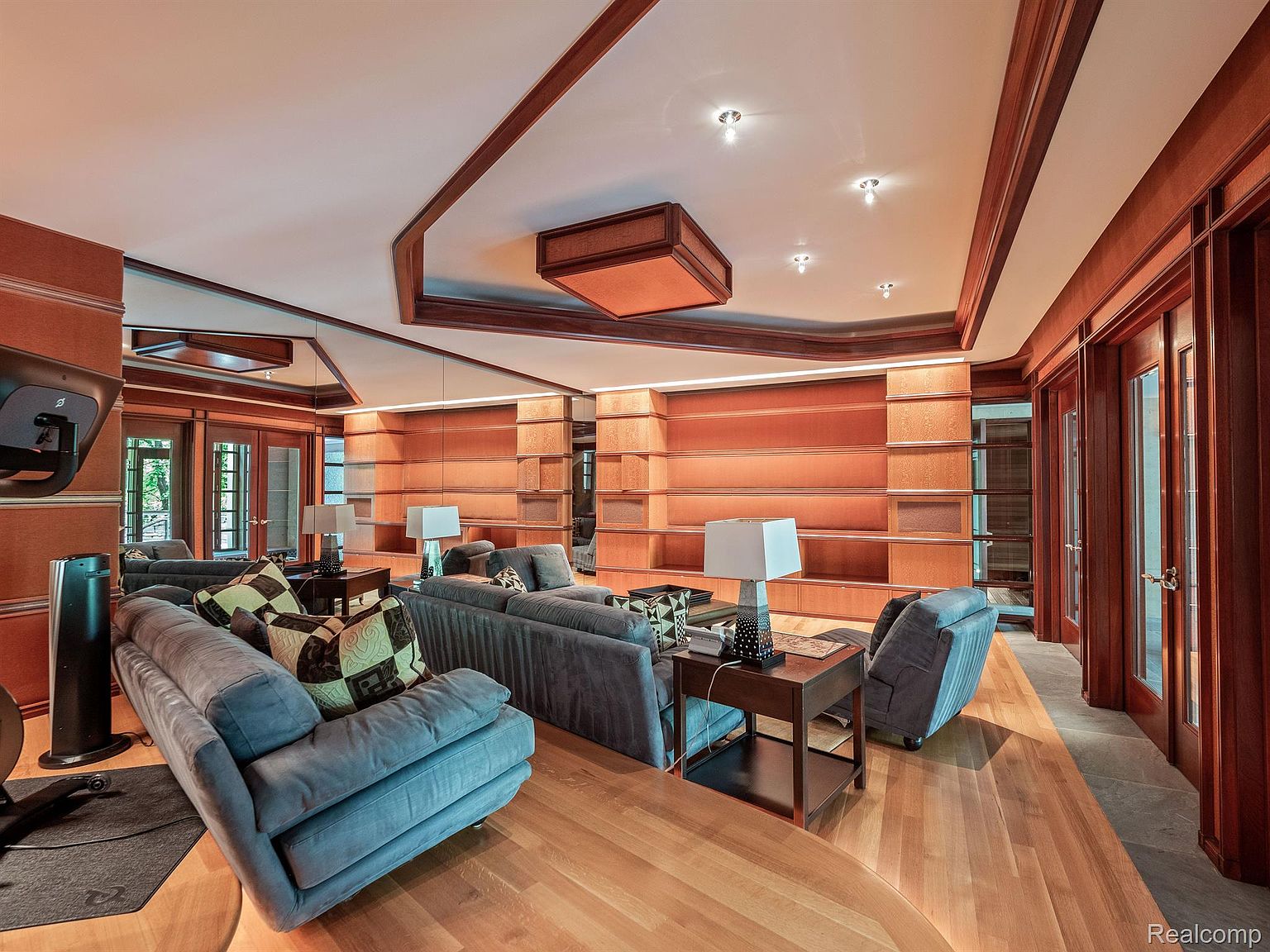 This is an interior shot of a living room featuring a large built-in bookshelf, two blue sofas, and a wooden floor. The room is well-lit with recessed lighting and table lamps, creating a warm and inviting atmosphere. The design incorporates wood trim and paneling, adding a touch of traditional elegance.
