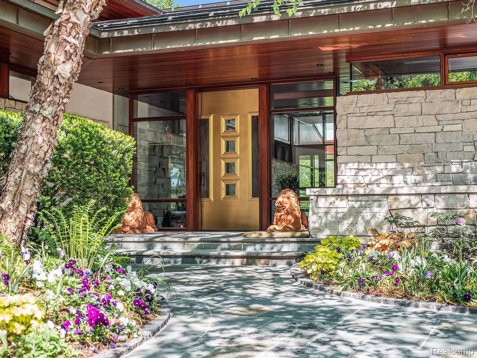 This is a captivating entryway to a home, featuring a striking gold door framed by glass panels and flanked by stone walls. Two lion statues guard the entrance, adding a touch of grandeur. The walkway is paved with stone, leading through a vibrant garden with colorful flowers, creating an inviting and luxurious first impression.