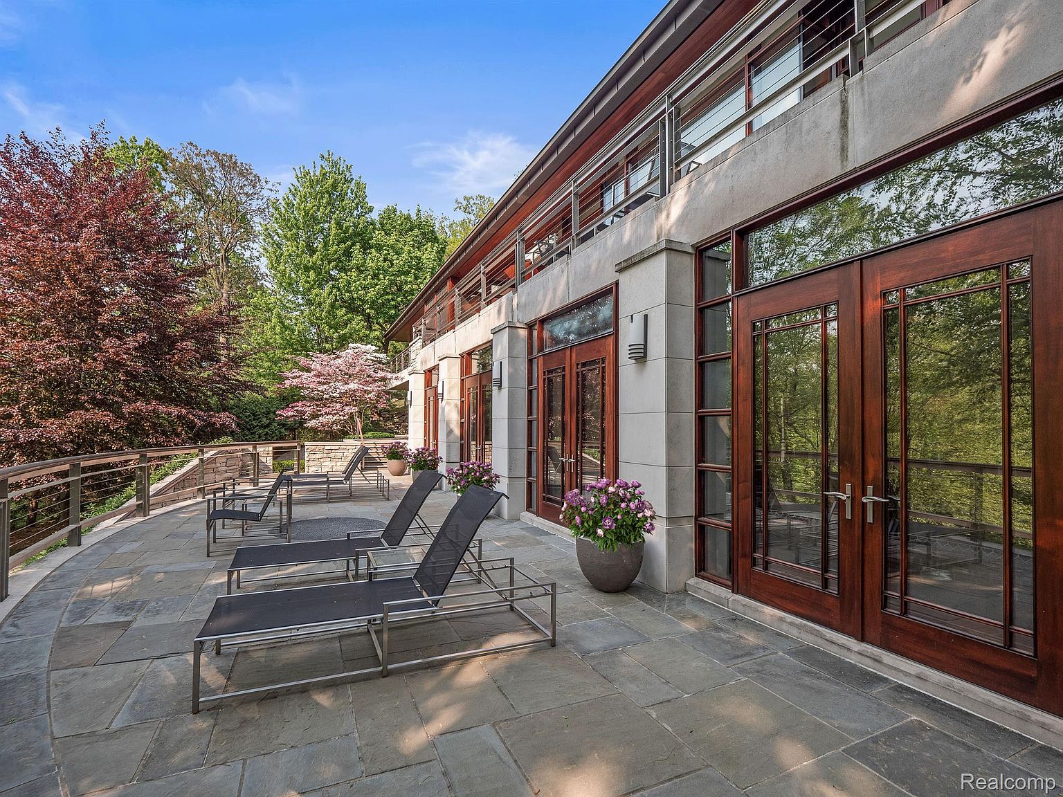 This image showcases a luxurious patio or balcony area, featuring sleek lounge chairs arranged on a stone-tiled surface. The space is bordered by a modern railing and offers views of lush greenery. Large, dark-framed glass doors provide access to the interior, suggesting a seamless indoor-outdoor living experience.