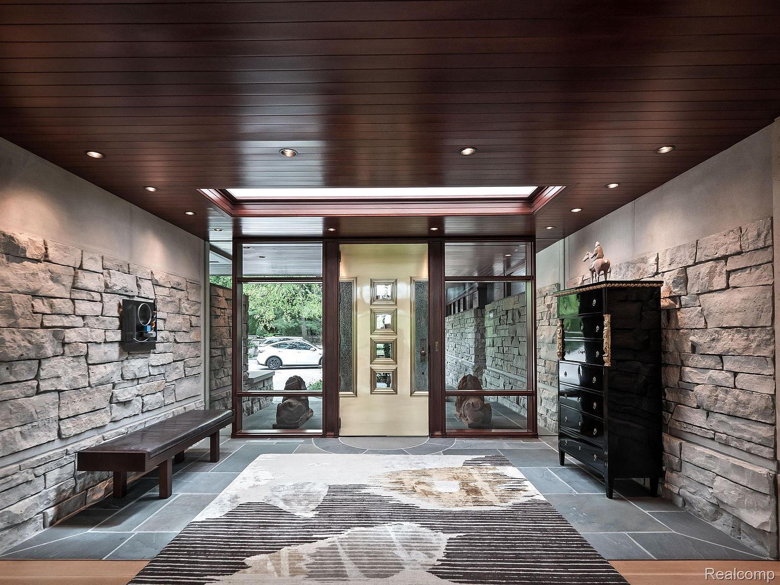 This interior shot showcases a grand entryway featuring stone walls, a dark wood ceiling, and a striking front door framed by large windows. A modern bench and a black chest of drawers add to the sophisticated ambiance, while a patterned rug anchors the space. The design blends natural elements with contemporary style, creating a welcoming and luxurious first impression.