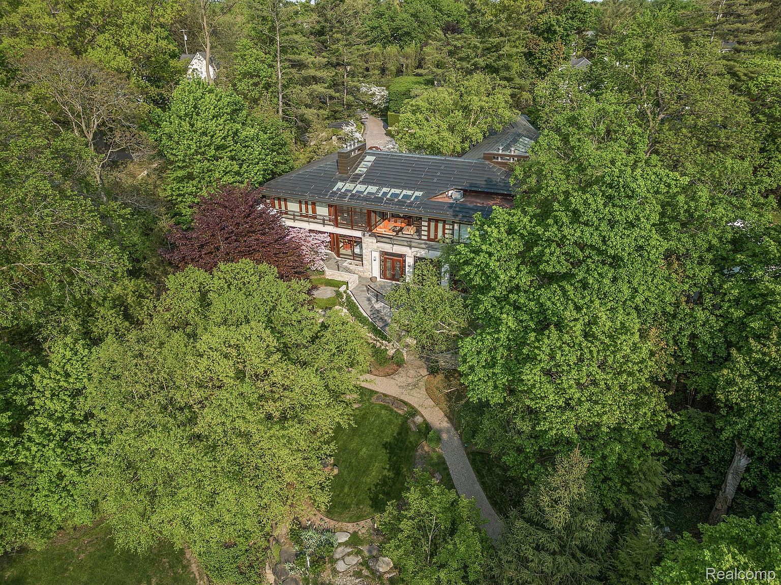 This aerial view showcases a luxurious home nestled among lush greenery. The house features a dark roof with skylights, a stone facade, and multiple windows and balconies. A winding stone pathway leads through the meticulously landscaped yard, highlighting the property's privacy and serene setting.