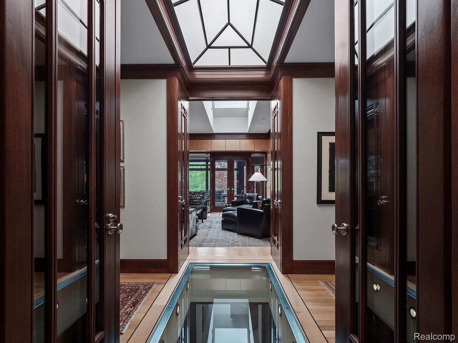 This interior shot showcases a luxurious hallway with rich wood trim and doors, leading to a living area with comfortable seating. A unique glass floor section provides a view to the level below, adding a modern architectural element. The space is well-lit with natural light from above, creating an inviting and sophisticated atmosphere.