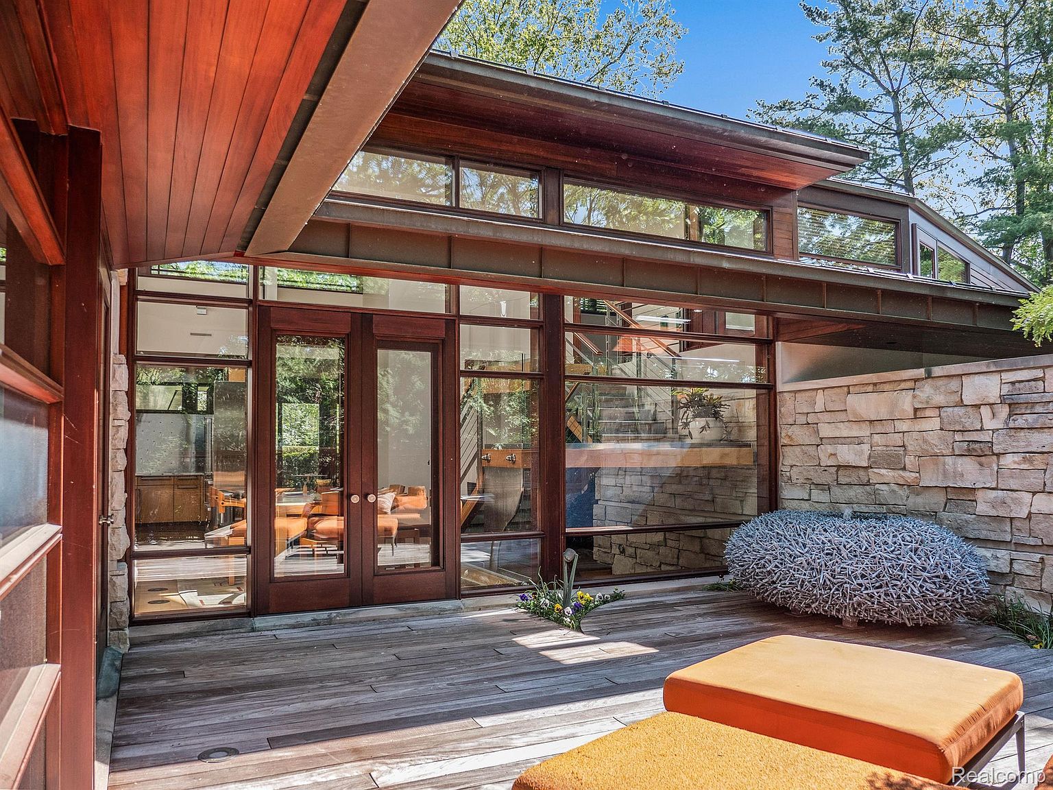 This image showcases a modern home's outdoor patio area, featuring a wooden deck, large glass windows and doors, and a stone accent wall. The patio includes unique decorative elements and comfortable seating, creating an inviting and stylish outdoor living space. The architecture emphasizes natural light and seamless integration with the surrounding landscape.