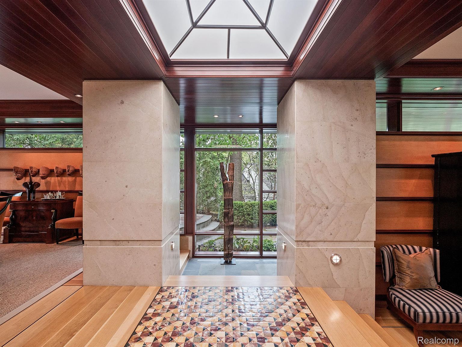 This interior shot showcases a hallway with unique architectural details, including stone pillars, a geometric skylight, and a patterned tile floor. A large window at the end of the hallway provides a view of the exterior greenery, creating a sense of depth and connection to nature. The space is well-lit and exudes a sophisticated, modern aesthetic.