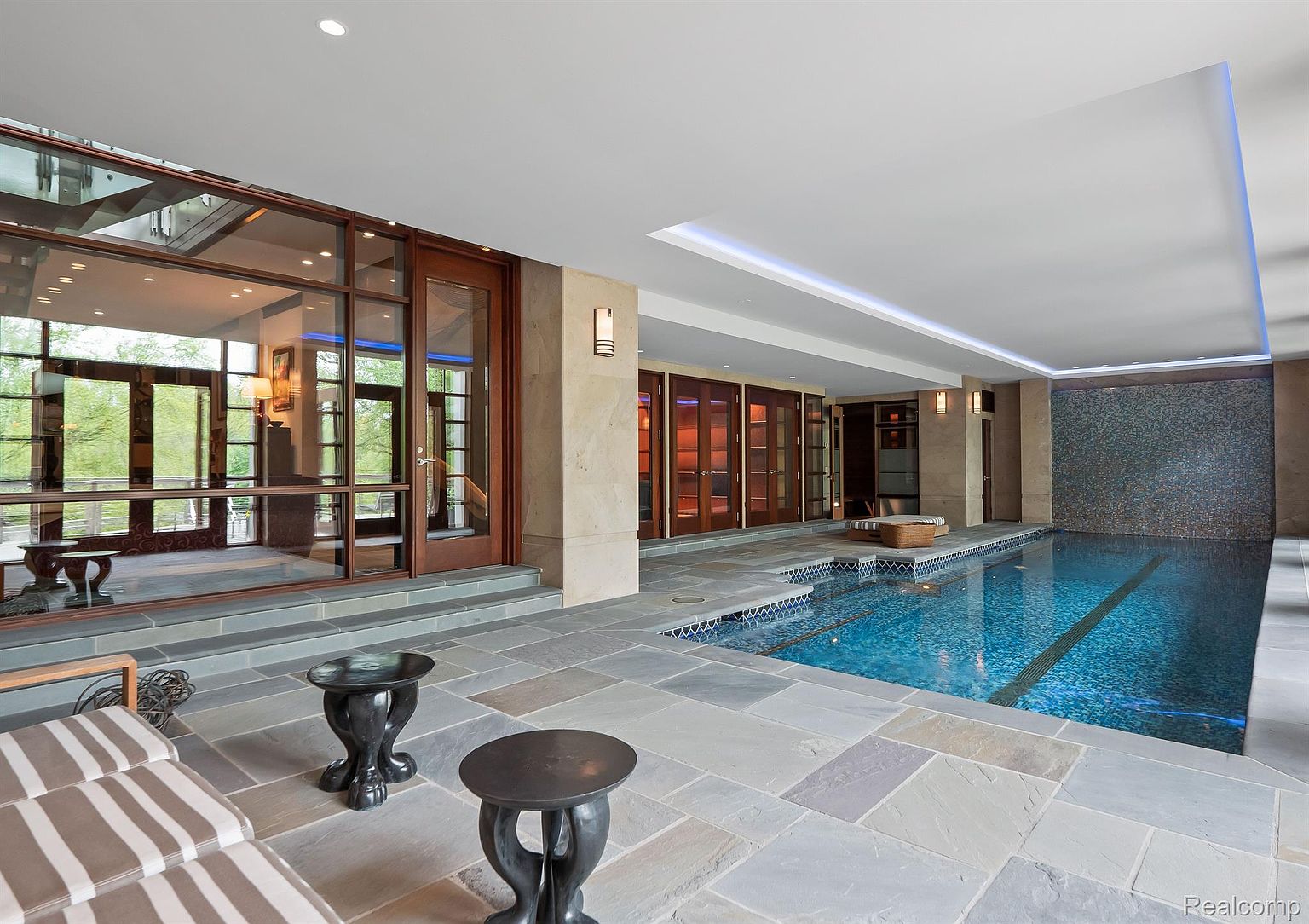 This image showcases an indoor pool area with a modern and luxurious design. The pool is surrounded by stone tile flooring and features a mosaic tile accent wall. Large windows and doors provide natural light and access to an outdoor area, while recessed lighting and wall sconces create a warm ambiance. The scene is captured from a low angle, emphasizing the depth and spaciousness of the pool area.