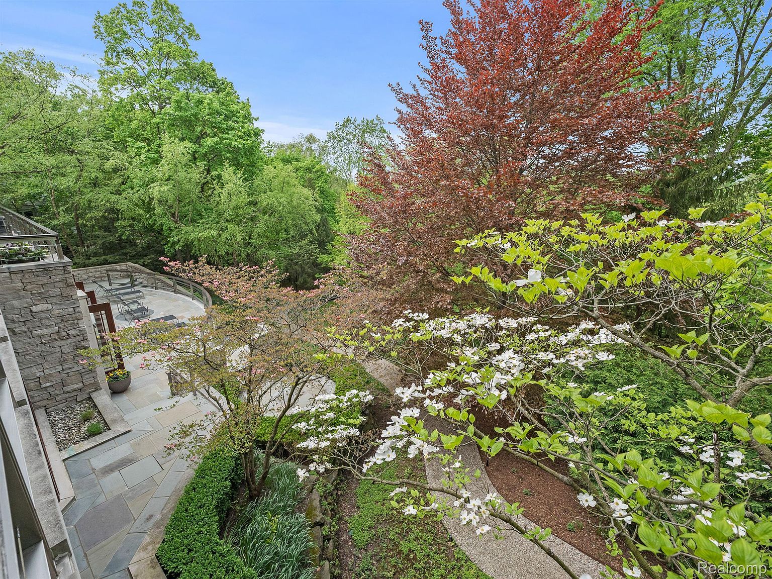 This image showcases a beautifully landscaped yard and garden area, featuring a stone patio with outdoor seating. Lush greenery, including flowering trees and manicured hedges, surrounds a winding pathway. The scene offers a serene and inviting outdoor living space, perfect for relaxation and entertaining.