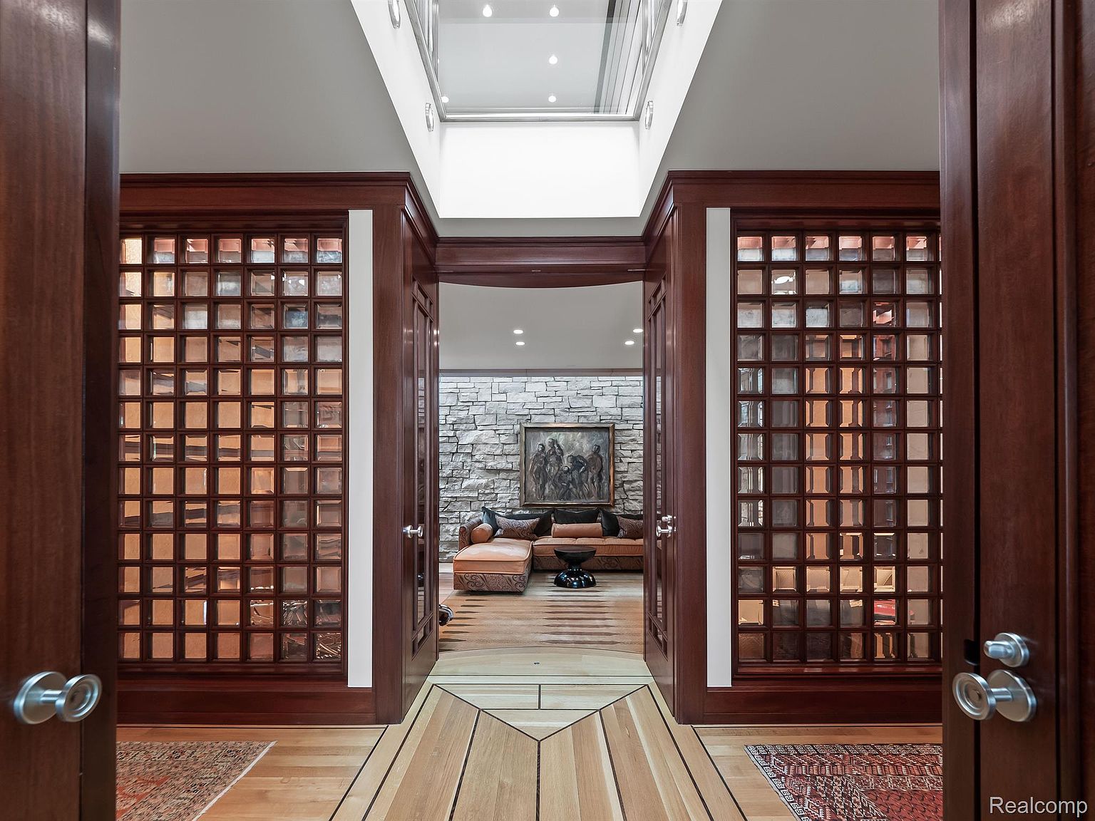 The image showcases an elegant entryway or hallway featuring a unique design with wooden framed glass block walls on either side. The flooring is a patterned light wood, leading into a living area with a stone accent wall and comfortable seating. A skylight provides natural light, enhancing the warm and inviting atmosphere.