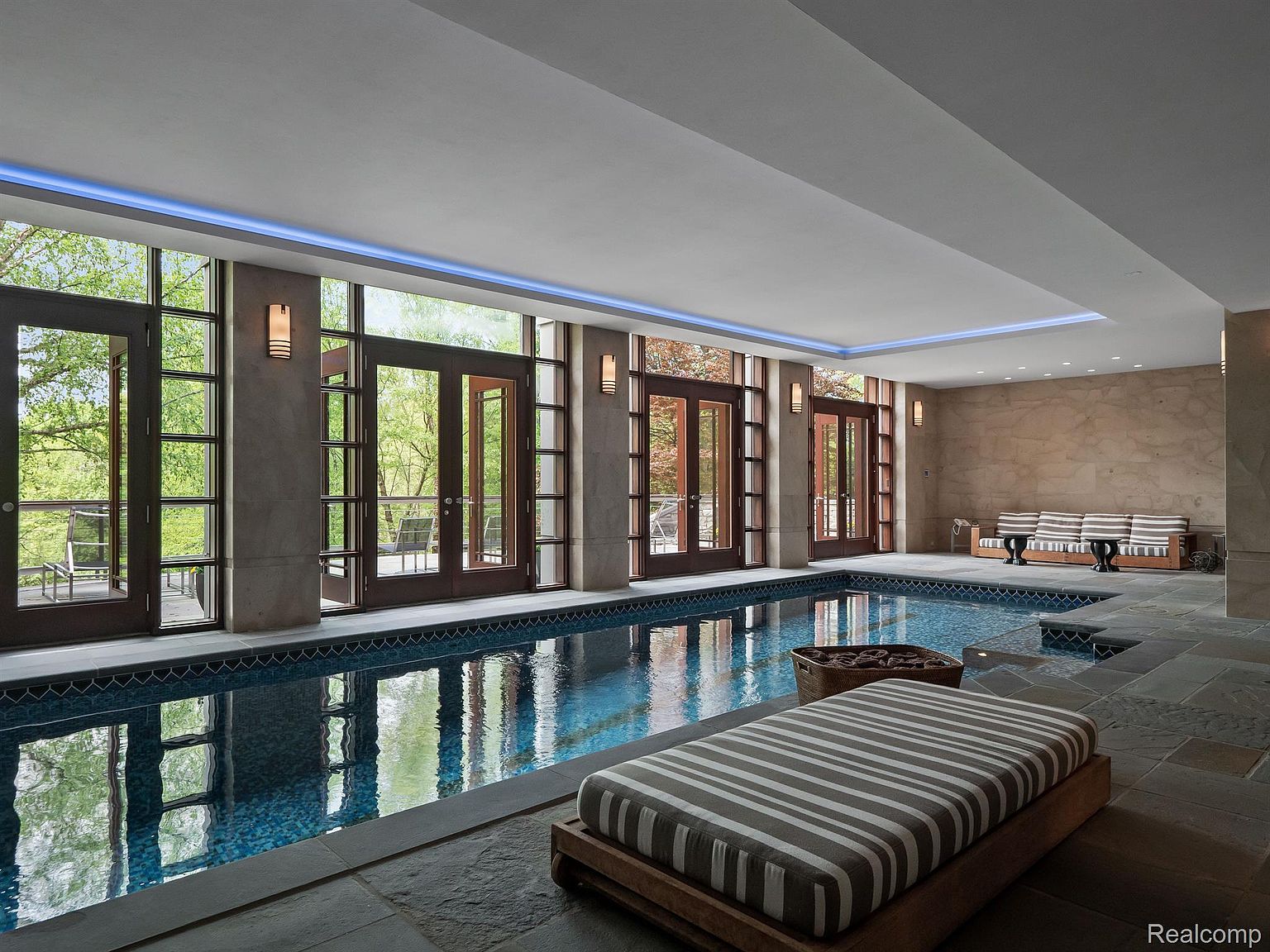 This image showcases a luxurious indoor pool area, featuring a long, rectangular pool with blue mosaic tiles. Large windows and doors line one side, offering views of the outdoors and allowing natural light to flood the space. A striped lounge chair sits poolside, and a seating area with striped cushions is visible in the background, creating an inviting and relaxing atmosphere.