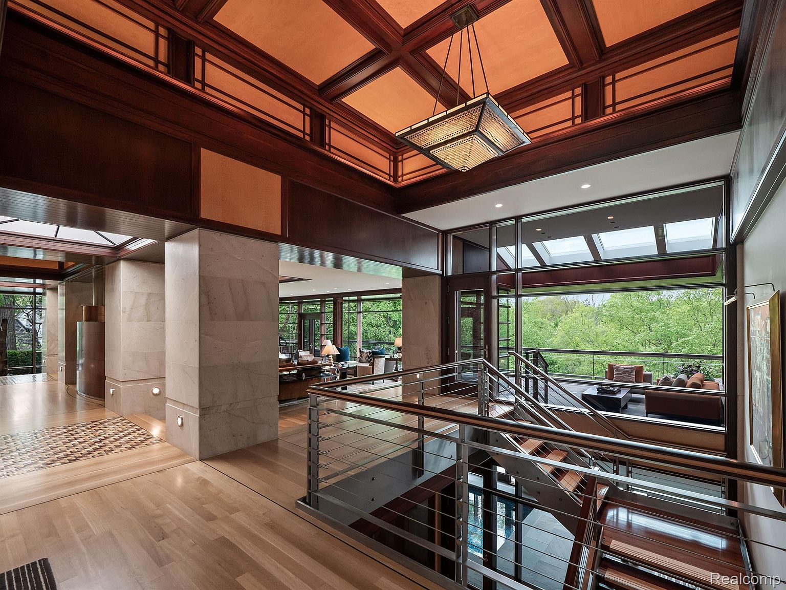 This interior shot showcases a luxurious hallway and staircase area. The space features a blend of natural wood flooring, marble columns, and a modern metal railing system. Large windows provide ample natural light and views of the surrounding greenery, enhancing the open and airy feel of the home.