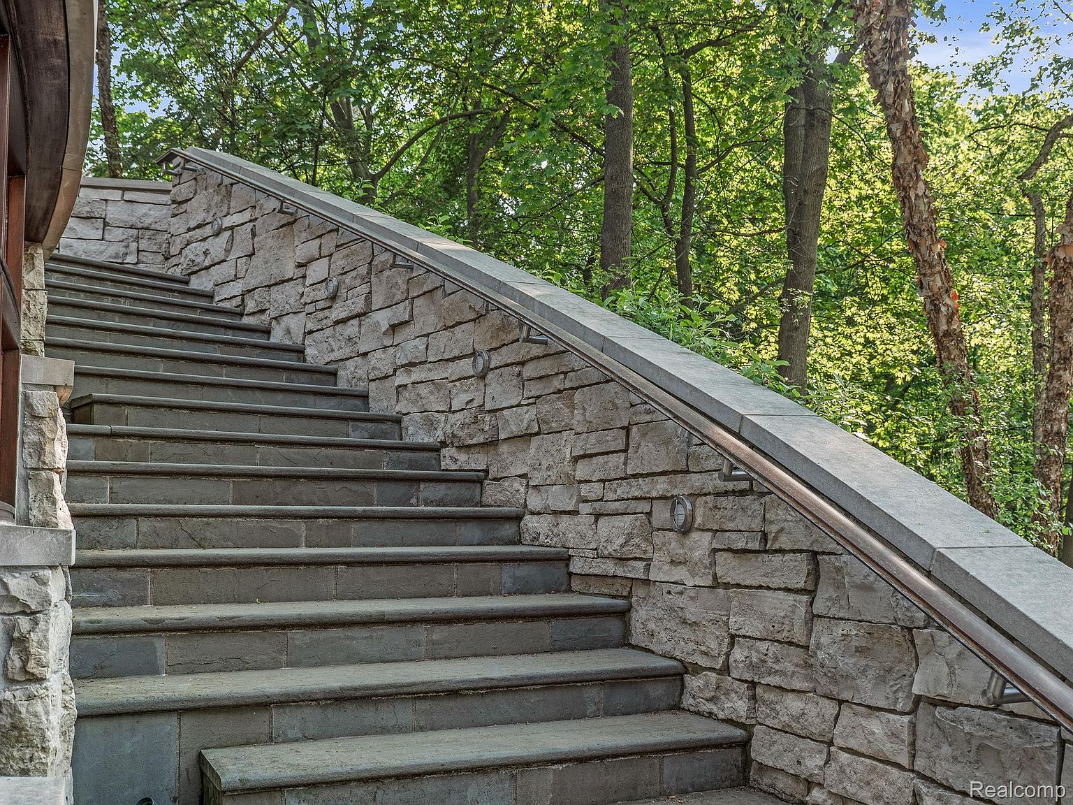 The image showcases a stone staircase leading upwards, possibly to a garden or another level of the property. The steps are constructed from gray stone, complemented by a retaining wall made of stacked, light-colored stones. A metal handrail runs along the wall, and the surrounding area is filled with lush greenery and trees, creating a natural and inviting atmosphere.