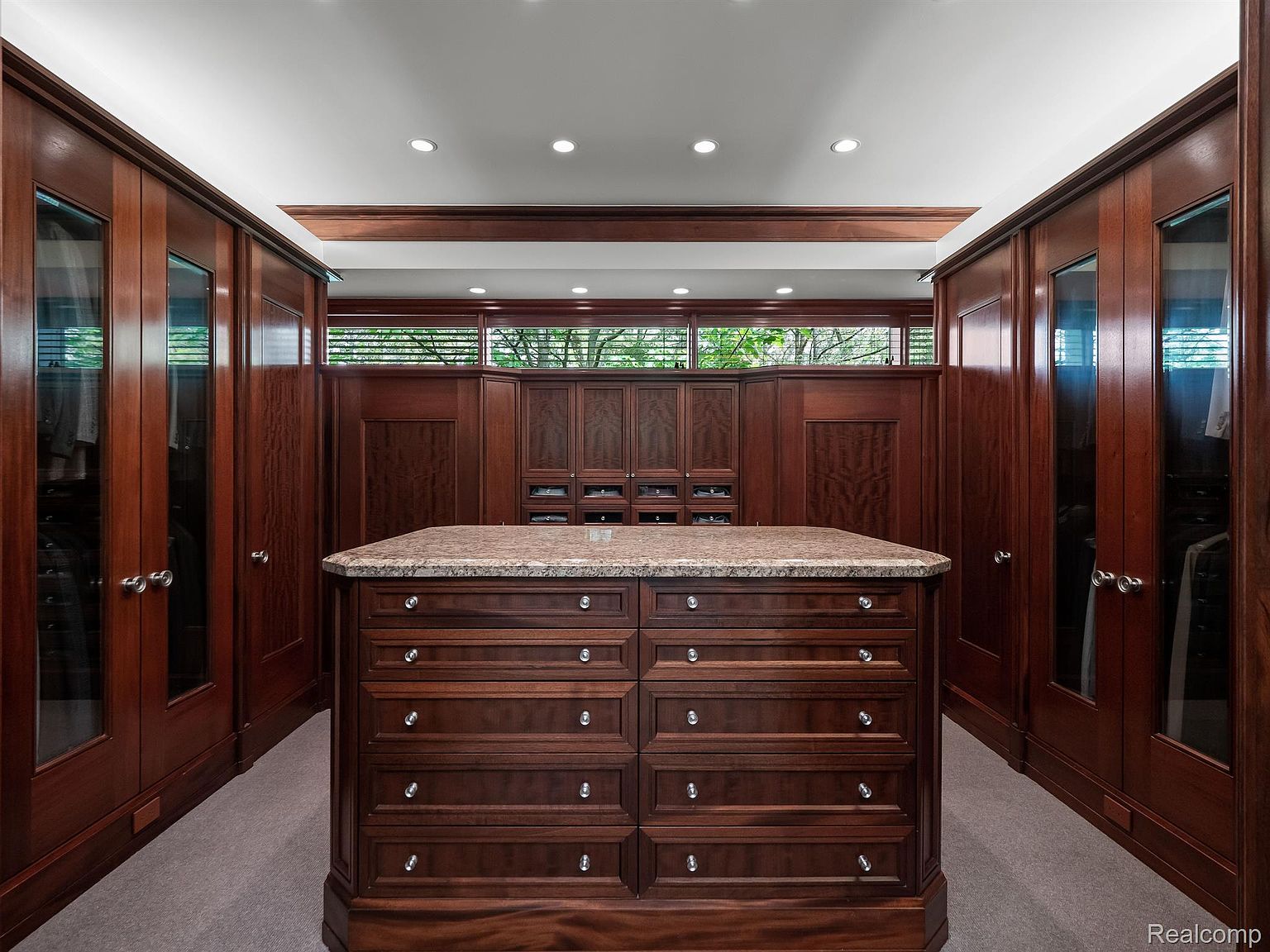 This is a well-organized walk-in closet featuring custom wood cabinetry and a central island with a granite countertop. The closet has ample storage space with drawers and hanging rods, and the design incorporates natural light through a high window. The overall impression is luxurious and functional.