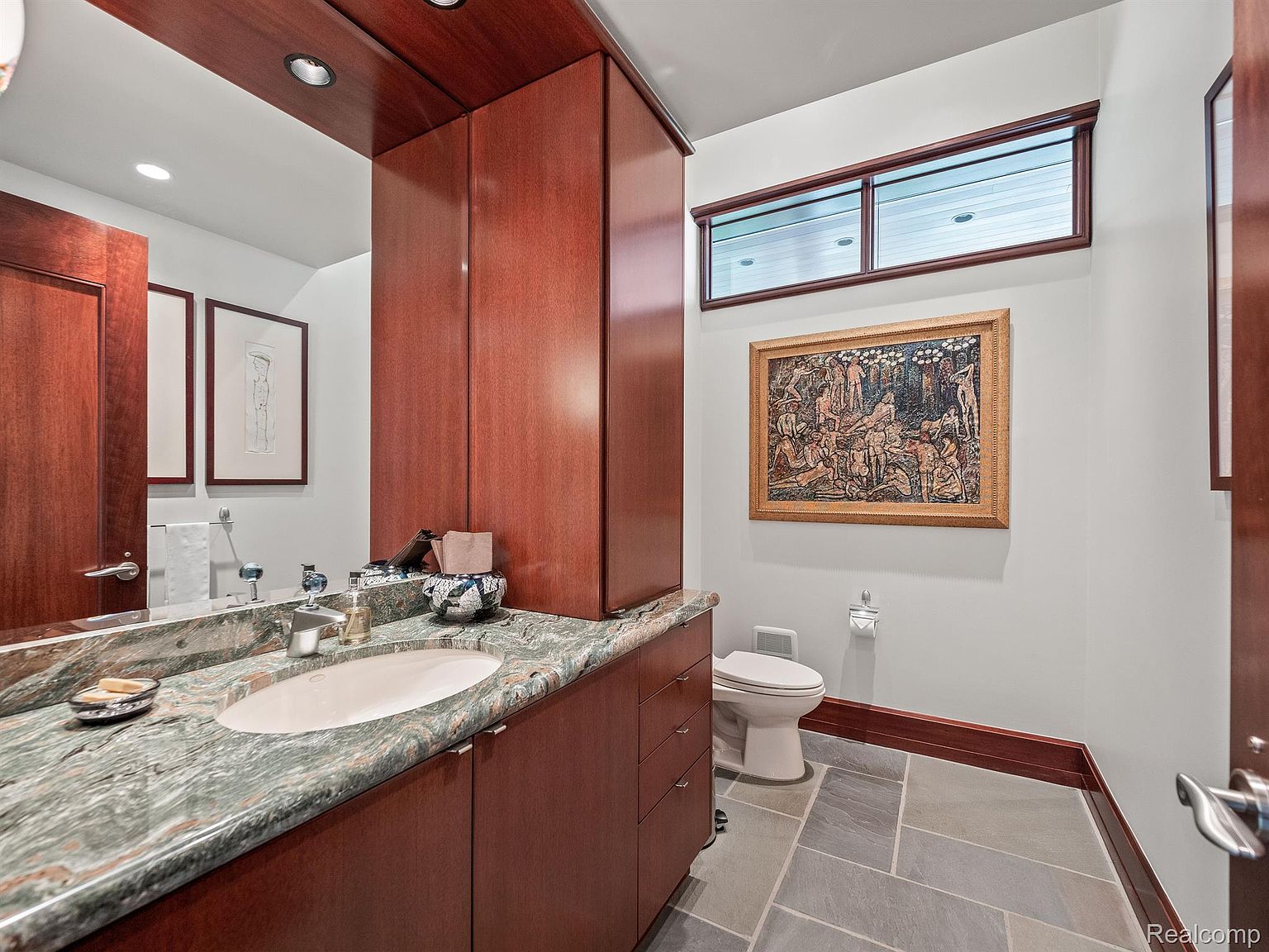 This bathroom features a unique green marble countertop with an oval sink, complemented by rich wood cabinetry. A framed artwork hangs above the toilet, and a small window provides natural light. The floor is tiled in a neutral gray, creating a sophisticated and clean aesthetic.