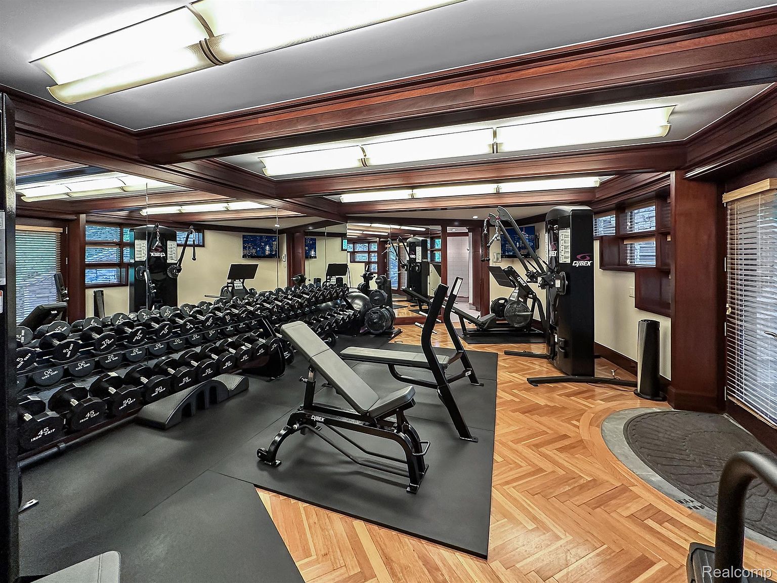 This is an interior shot of a well-equipped home gym. The space features a variety of weight training equipment, including dumbbells, benches, and Cybex machines. The flooring transitions from black rubber mats to a herringbone wood pattern, and the room is well-lit with recessed lighting and natural light from the windows.