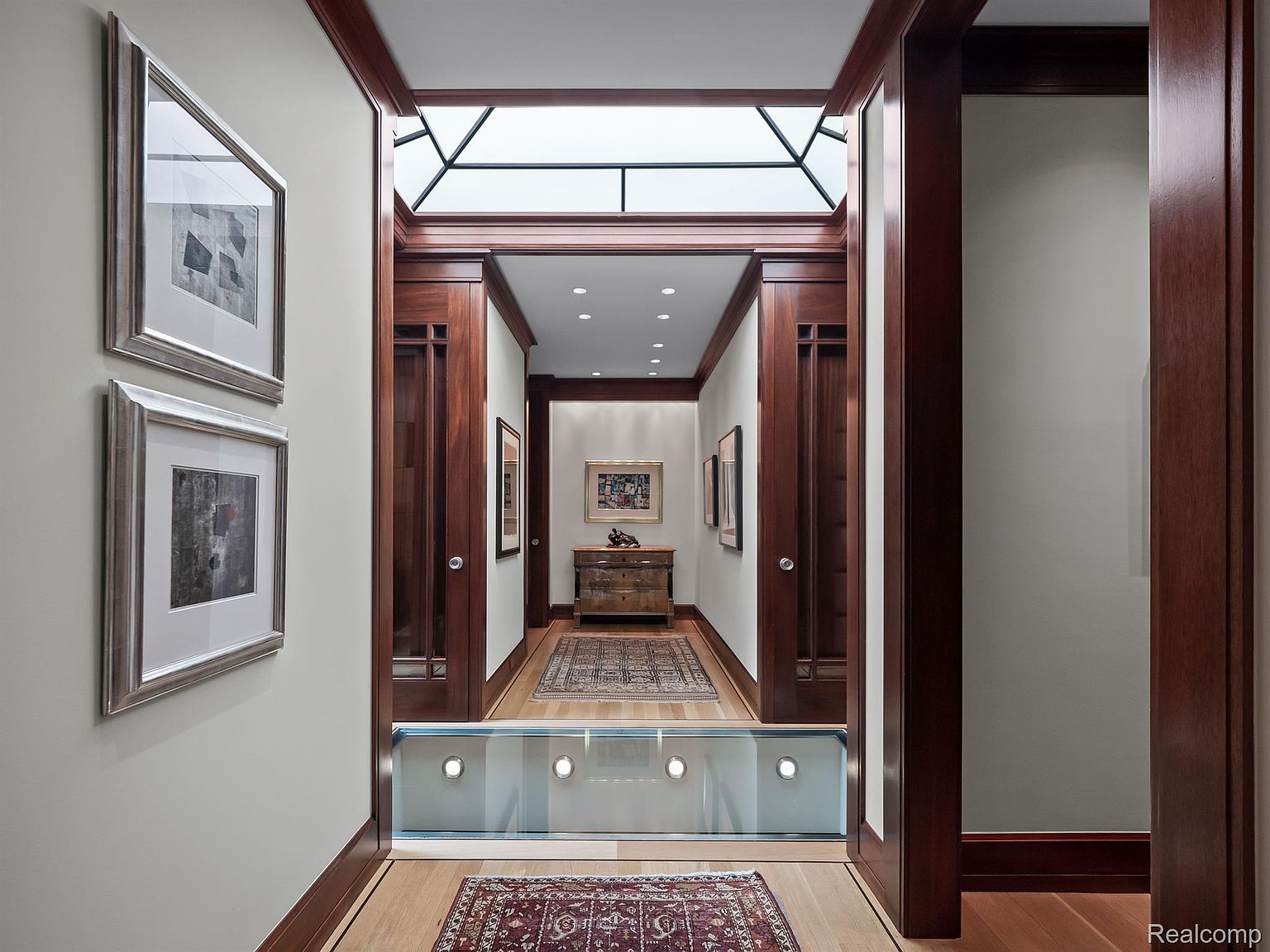 This interior shot showcases a luxurious hallway with rich wood trim and a unique glass-enclosed lower level, illuminated by recessed lighting. Artwork adorns the walls, and a skylight provides natural light, enhancing the elegant and sophisticated ambiance. The hallway leads to a well-appointed space with a chest of drawers and additional artwork, suggesting a seamless flow within the home.