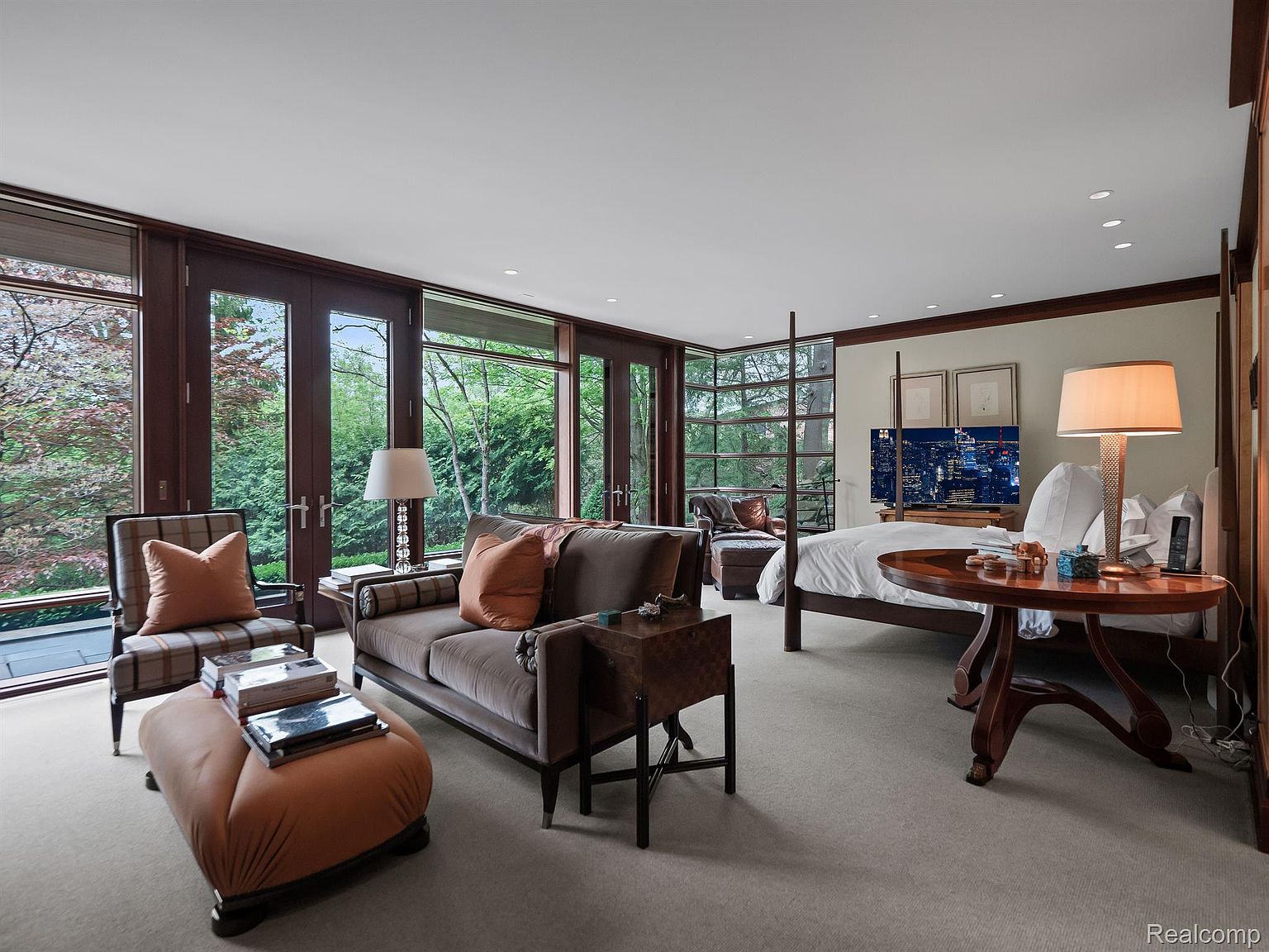 This is a luxurious primary bedroom featuring a four-poster bed with white linens, a sitting area with a brown sofa and armchair, and large windows offering views of lush greenery. The room is decorated in neutral tones with wood accents, creating a serene and inviting atmosphere. A round side table with a lamp sits next to the bed, and a television is mounted on a dresser.