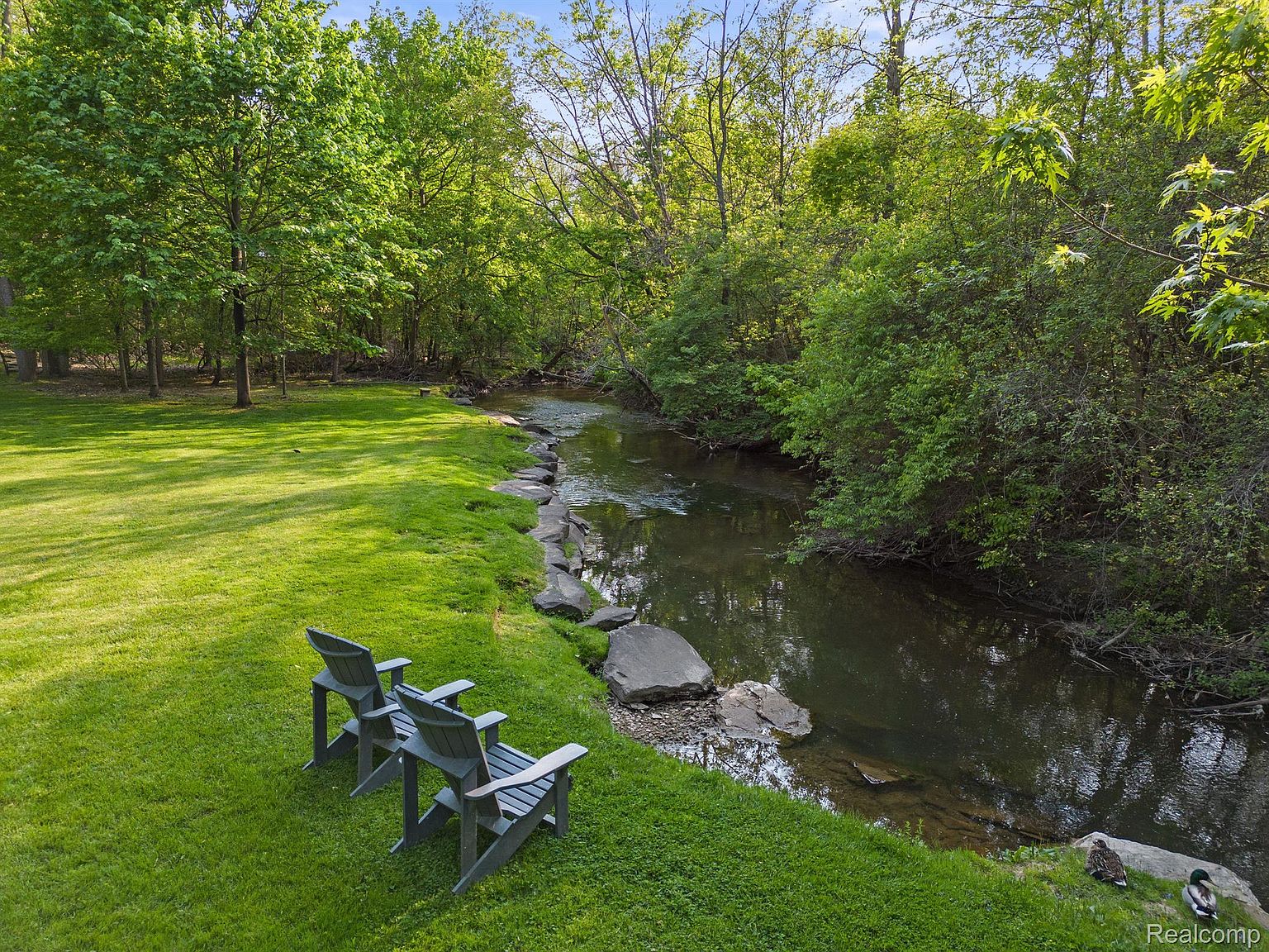 This exterior shot showcases a serene backyard featuring a lush green lawn that slopes gently towards a tranquil stream. Two Adirondack chairs are placed on the grass, inviting relaxation by the water's edge. The scene is framed by mature trees and verdant foliage, creating a peaceful and private outdoor space.