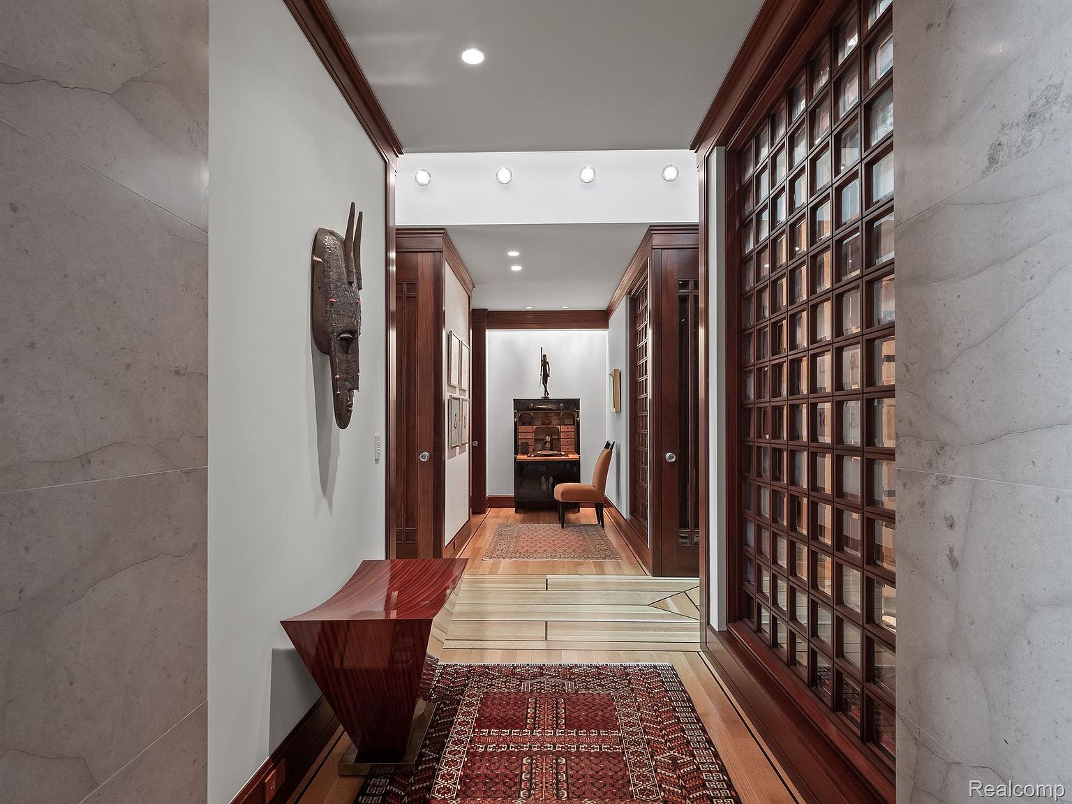 This interior shot showcases a well-appointed hallway featuring rich wood trim, a decorative wall mask, and a stylish console table. The hallway leads to another room with a patterned rug and a glimpse of furniture, creating a sense of depth and luxury. The design elements suggest a sophisticated and elegant home.