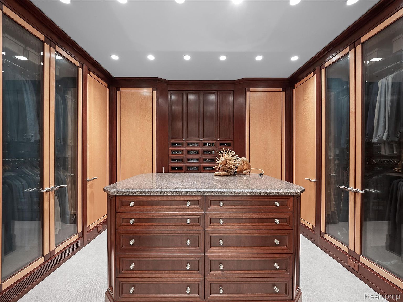 This is a well-organized walk-in closet featuring custom cabinetry with a rich, dark wood finish and light wood accents. The closet includes glass-fronted wardrobes and a central island with drawers and a granite countertop. Recessed lighting illuminates the space, creating a luxurious and functional dressing area.