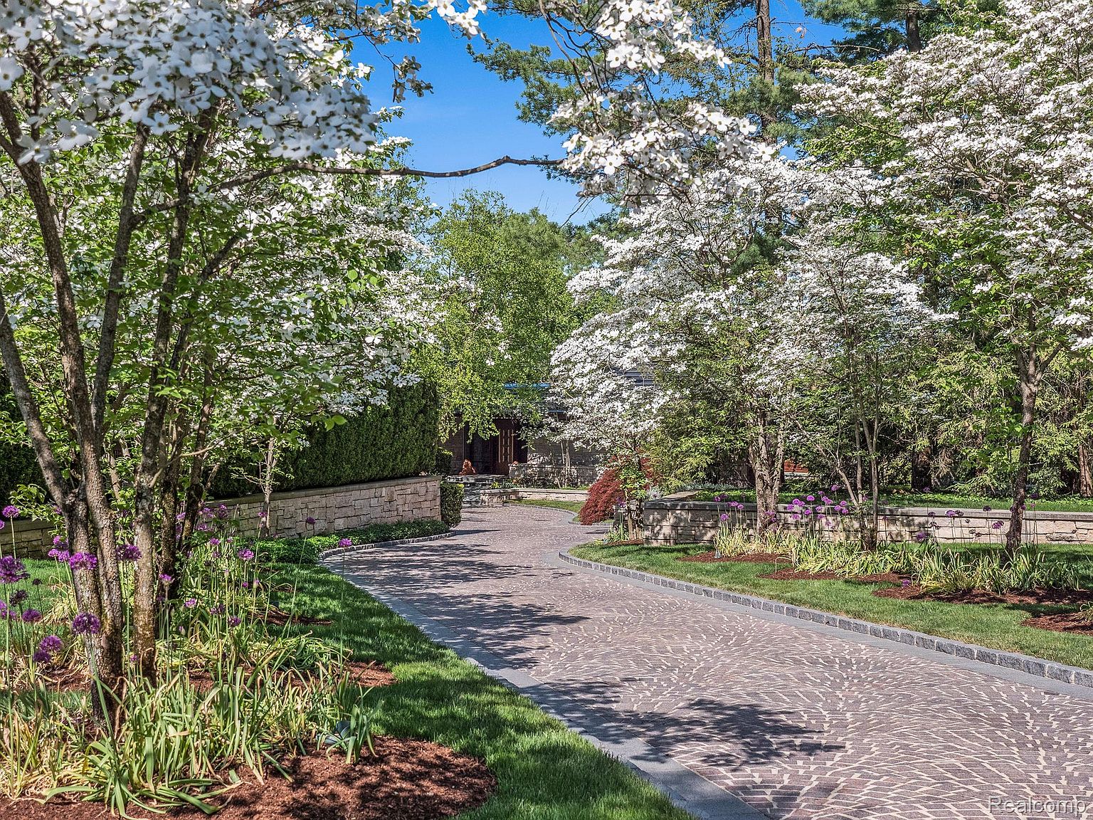 This image showcases a beautifully landscaped yard and garden, featuring a winding brick driveway lined with lush greenery, flowering trees, and vibrant purple allium flowers. A low stone wall adds architectural interest, while the overall impression is one of tranquility and meticulous care, enhancing the property's curb appeal. The perspective is from the entrance of the property, looking towards the house.