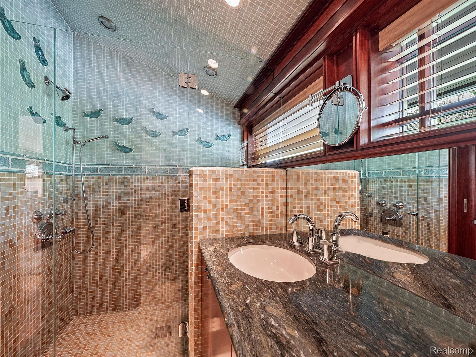 This is a primary bathroom featuring a double sink vanity with a dark granite countertop and a large mirror. The shower is enclosed with glass and features mosaic tile walls with decorative fish accents. The overall style is luxurious and unique, with a focus on high-end finishes and artistic details.
