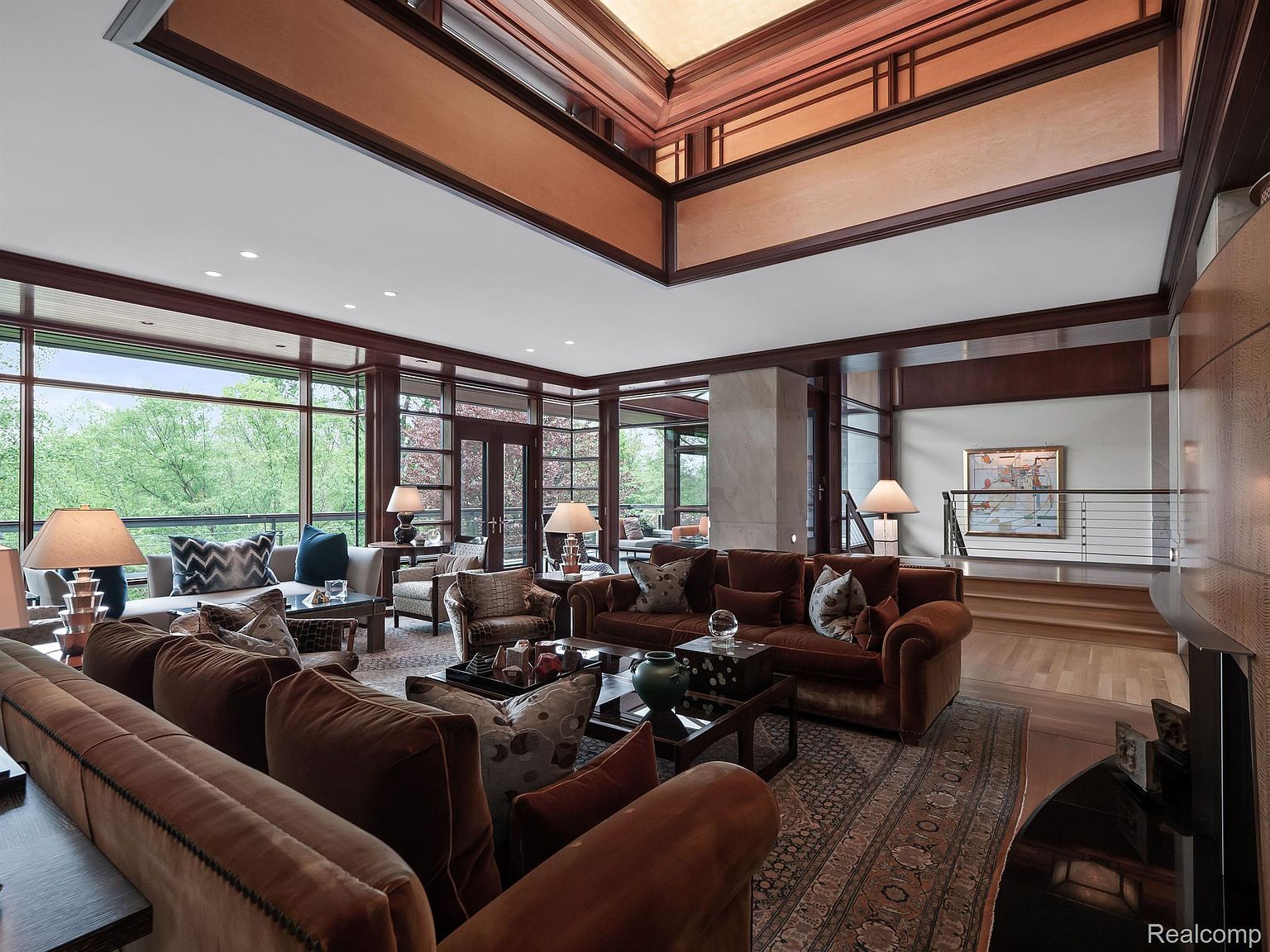 This is a spacious living room featuring a high, coffered ceiling with wood accents and large windows offering natural light and views of the surrounding greenery. The room is furnished with multiple brown sofas, patterned armchairs, and dark wood coffee tables, creating a warm and inviting atmosphere. An area rug anchors the seating arrangement, and a staircase is visible in the background, suggesting a multi-level home.