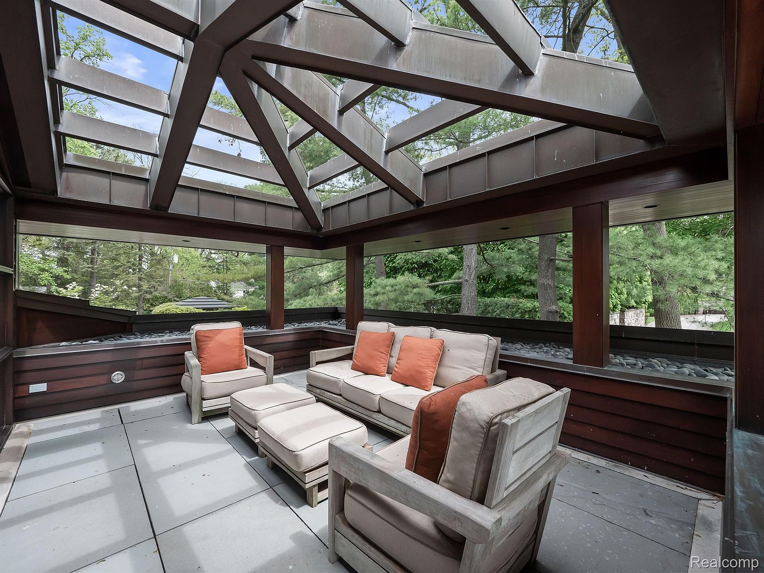 This sunroom features a modern design with a unique geometric skylight and large windows offering views of the surrounding greenery. The seating area includes a sofa, chairs, and ottomans, all upholstered in a neutral fabric with orange accent pillows. The flooring is tiled, and the room is decorated with dark wood accents and a pebble-filled ledge, creating a serene and inviting atmosphere.