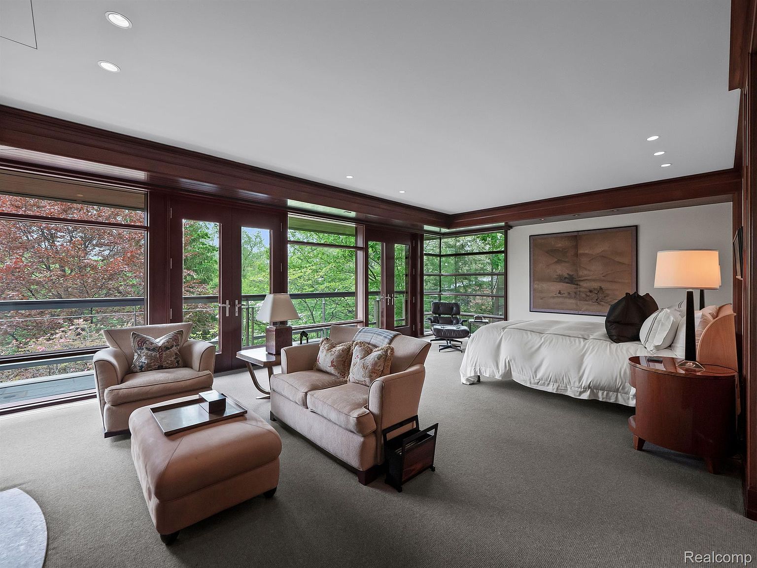 This is a spacious primary bedroom featuring a seating area with a sofa, armchair, and ottoman, as well as a large bed with white linens. The room has dark wood trim around the windows and ceiling, and a large piece of artwork hangs above the bed. Large windows offer views of the surrounding greenery, creating a serene and luxurious atmosphere.