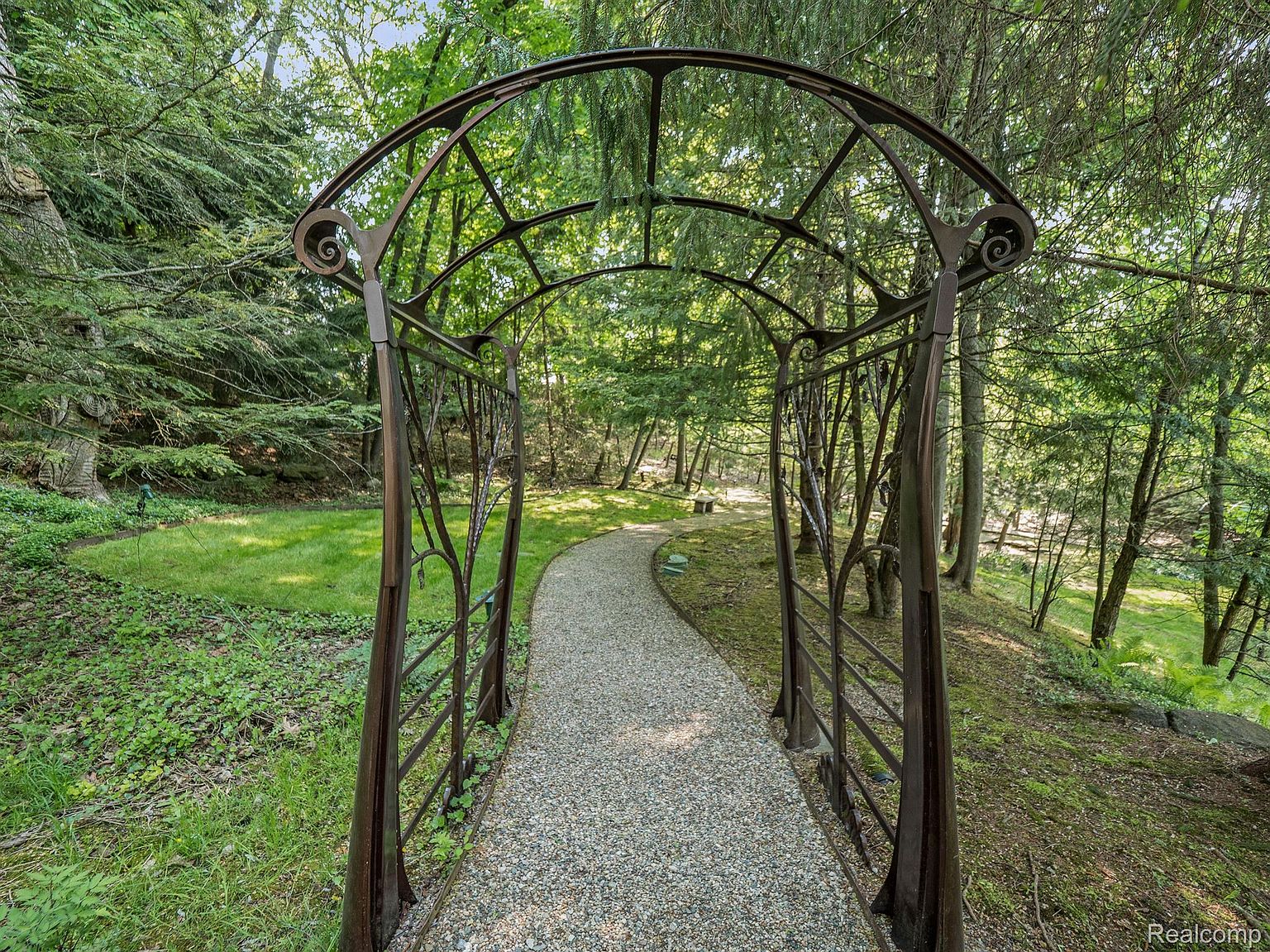 The image showcases a charming garden pathway framed by an ornate metal archway. The path is covered in gravel and winds through a lush green landscape with mature trees and foliage. The scene evokes a sense of tranquility and natural beauty, highlighting the property's outdoor space.