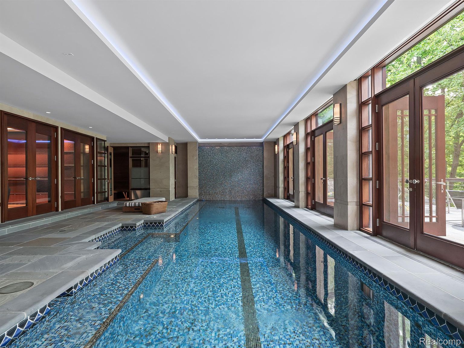 This image showcases a luxurious indoor swimming pool. The pool features blue mosaic tiles and a dark lane marker, surrounded by gray stone tiling. Large wooden-framed glass doors line one side, providing natural light and access to the outdoors, while the opposite side features wooden doors and a mosaic tiled wall at the far end. The ceiling is white with recessed lighting, creating a bright and inviting space.