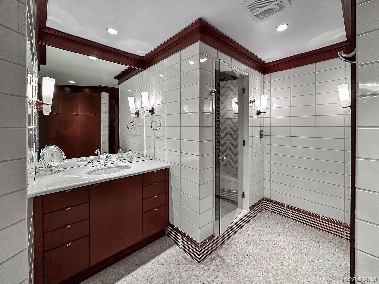 This is a well-lit primary bathroom featuring white tiled walls with a dark wood trim. A vanity with a white countertop and dark wood cabinets is visible, along with a large mirror and sconce lighting. A glass-enclosed shower with patterned tile inside is also present, contributing to a clean and modern aesthetic.