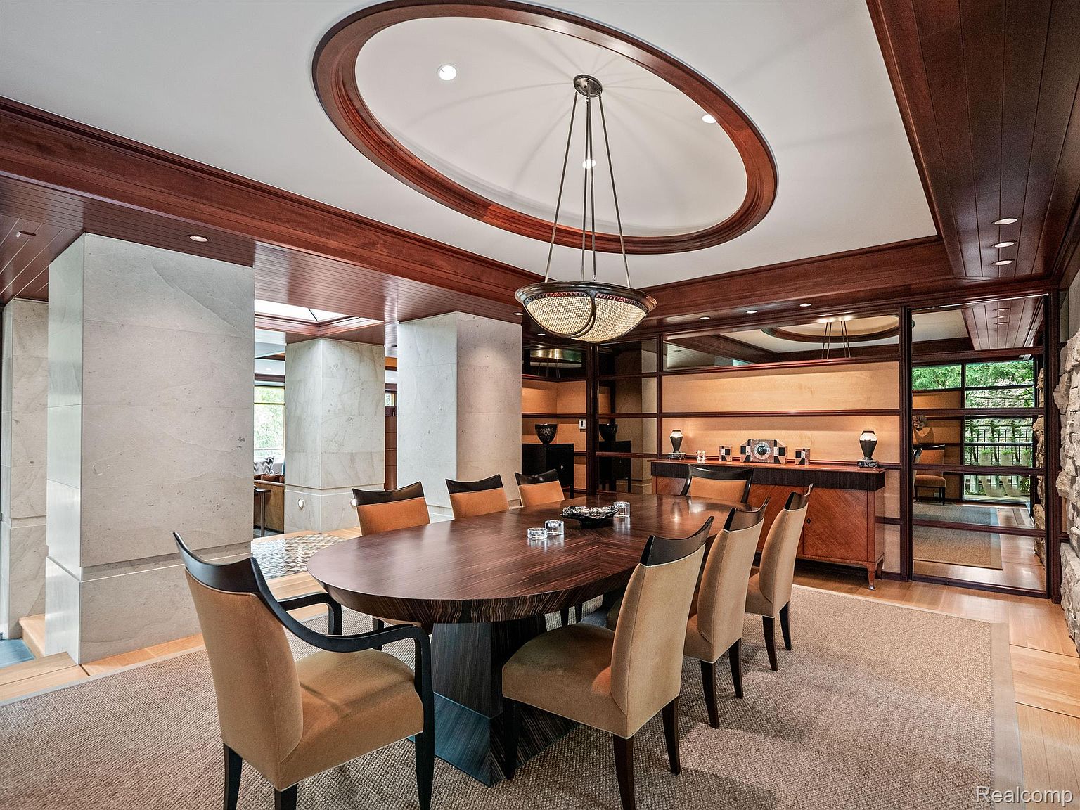 This is an interior shot of a dining room featuring a large, oval wooden table surrounded by upholstered chairs with dark wood accents. A modern chandelier hangs above the table, and the room is decorated with wood paneling and built-in shelving. The space is well-lit and appears luxurious, with a neutral-toned rug grounding the setting.