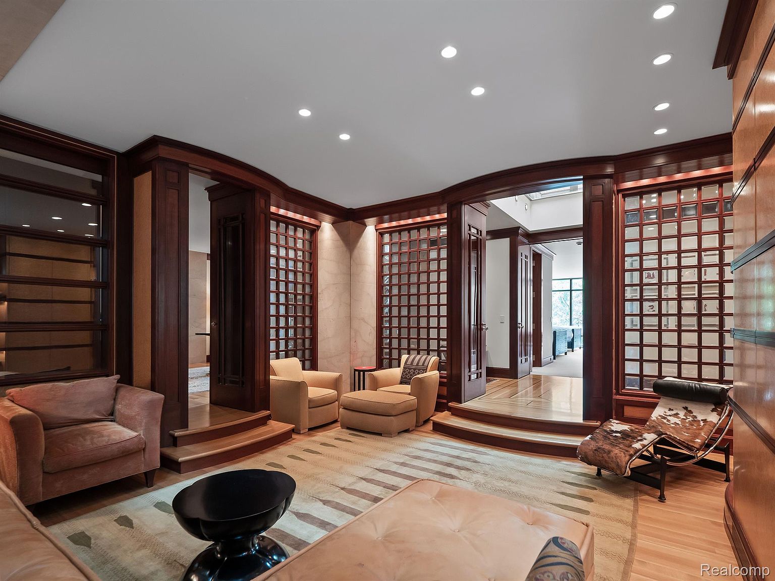 This is an interior shot of a luxurious living room featuring rich wood paneling and trim, creating a warm and sophisticated ambiance. The room is furnished with comfortable seating arrangements, including armchairs and a plush ottoman, arranged around a stylish coffee table. The design incorporates architectural details such as stepped platforms and grid-patterned walls, adding depth and visual interest to the space.