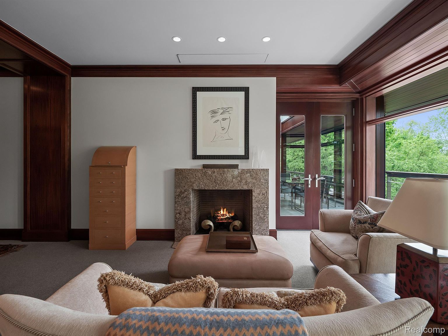 This is an interior shot of a living room featuring a fireplace with a stone surround and a framed piece of art above it. The room is furnished with comfortable seating, including a sofa and armchair, and a large ottoman. The space is well-lit with recessed lighting and natural light coming through a large window, creating a warm and inviting atmosphere.