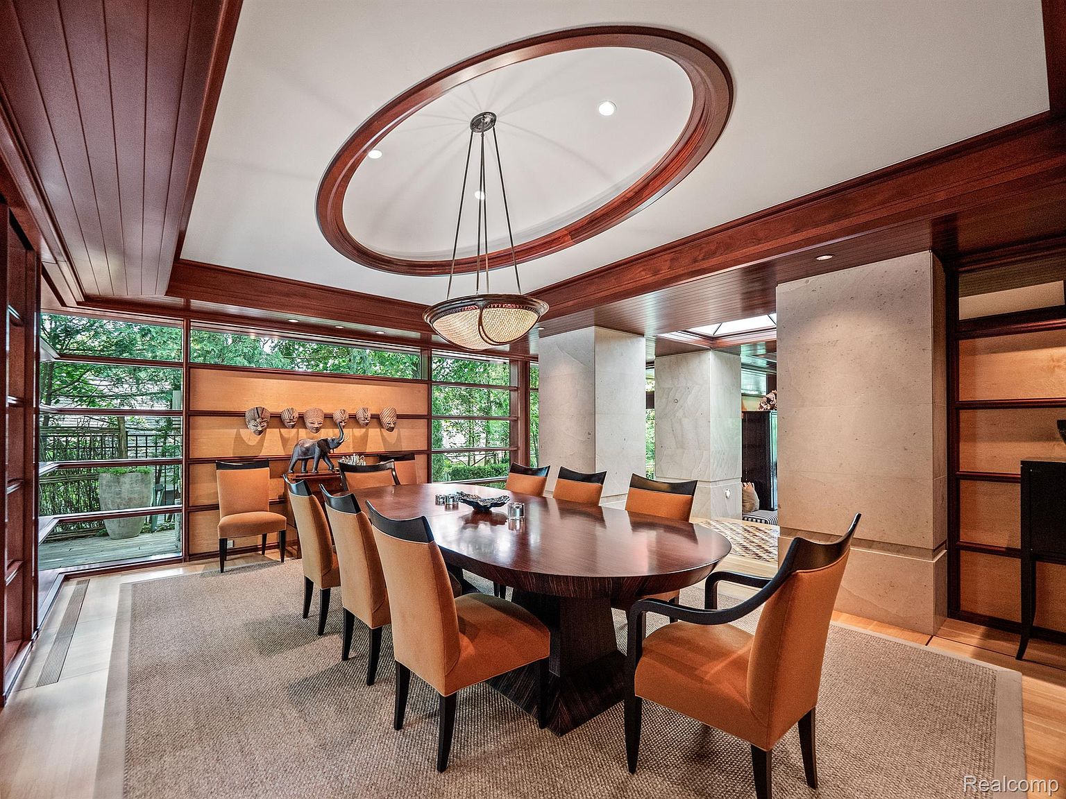 This is a dining room featuring a large oval wooden table surrounded by orange upholstered chairs with black frames. The room has a modern design with wood paneling, large windows offering natural light, and a unique oval ceiling feature with a chandelier. The space is elegant and well-lit, creating a welcoming atmosphere for dining.
