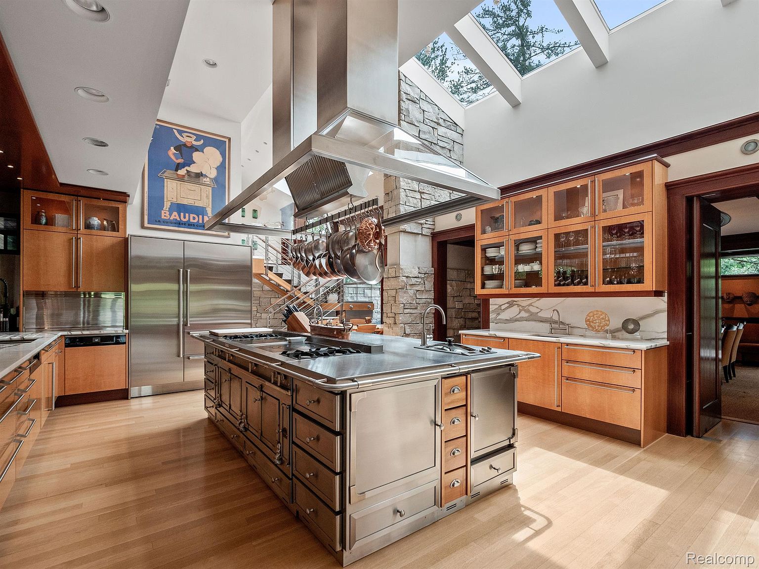 This is a spacious, high-end kitchen featuring a large stainless steel island with a built-in cooktop and ample storage. The kitchen boasts custom wood cabinetry, stainless steel appliances, and a unique stone accent wall with a skylight above. The hardwood floors and natural light create a warm and inviting atmosphere.