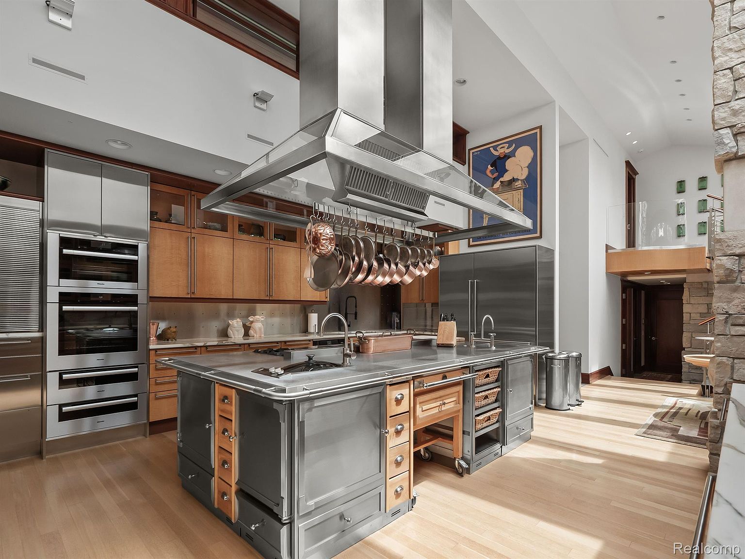 This is a spacious, high-end kitchen featuring a large central island with a stainless steel countertop and custom cabinetry. The kitchen is equipped with stainless steel appliances, including double ovens and a refrigerator, and a prominent overhead vent hood with hanging copper pots. The open layout and light wood flooring create a bright and inviting atmosphere.
