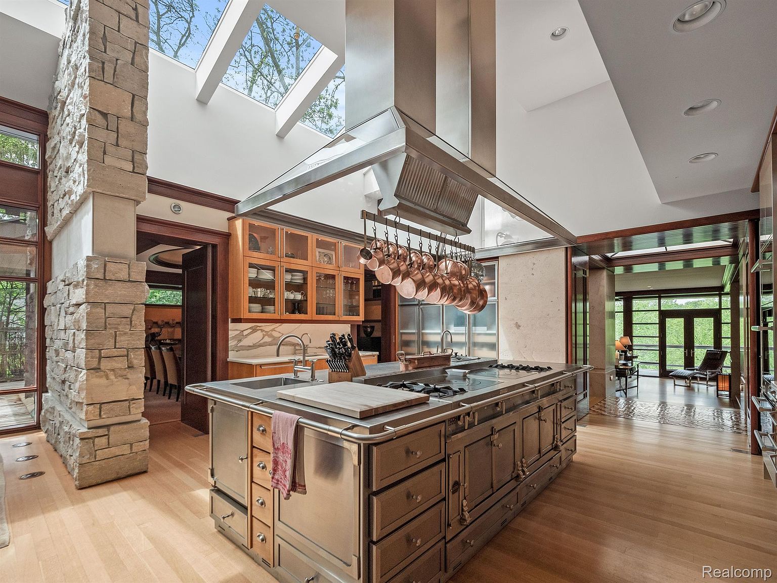 This is a spacious, high-end kitchen featuring a large stainless steel island with a built-in cooktop and ample storage. The kitchen boasts a unique design with a skylight, a stone pillar, and copper pots hanging above the island. The hardwood floors and natural wood cabinetry add warmth to the space, creating a luxurious and functional culinary environment.