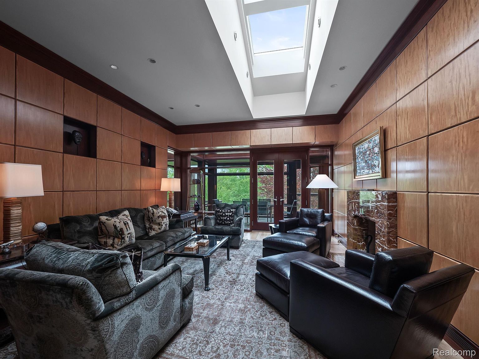 This is an interior shot of a luxurious living room featuring wood-paneled walls, a skylight, and a marble fireplace. The room is furnished with plush sofas, leather armchairs, and a glass-topped coffee table, all arranged on an ornate rug. The perspective is from a medium angle, showcasing the room's depth and the natural light streaming in from above.